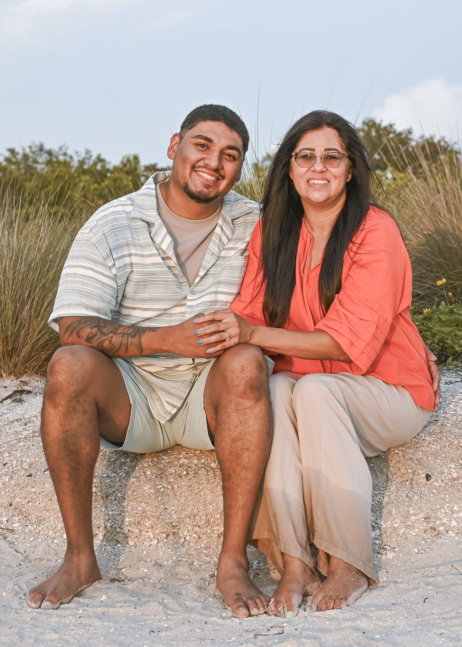 A smiling couple sits together on a sandy beach, the man with a tattoo wearing a striped shirt and the woman in coral top.