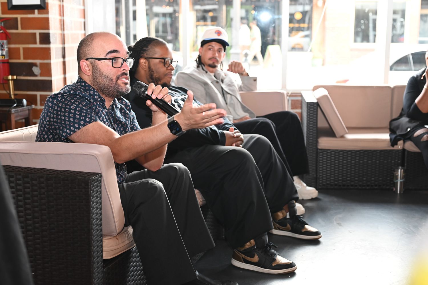 Three men seated on lounge chairs during a panel discussion, one in a plaid shirt and glasses speaking into a microphone and gesturing.