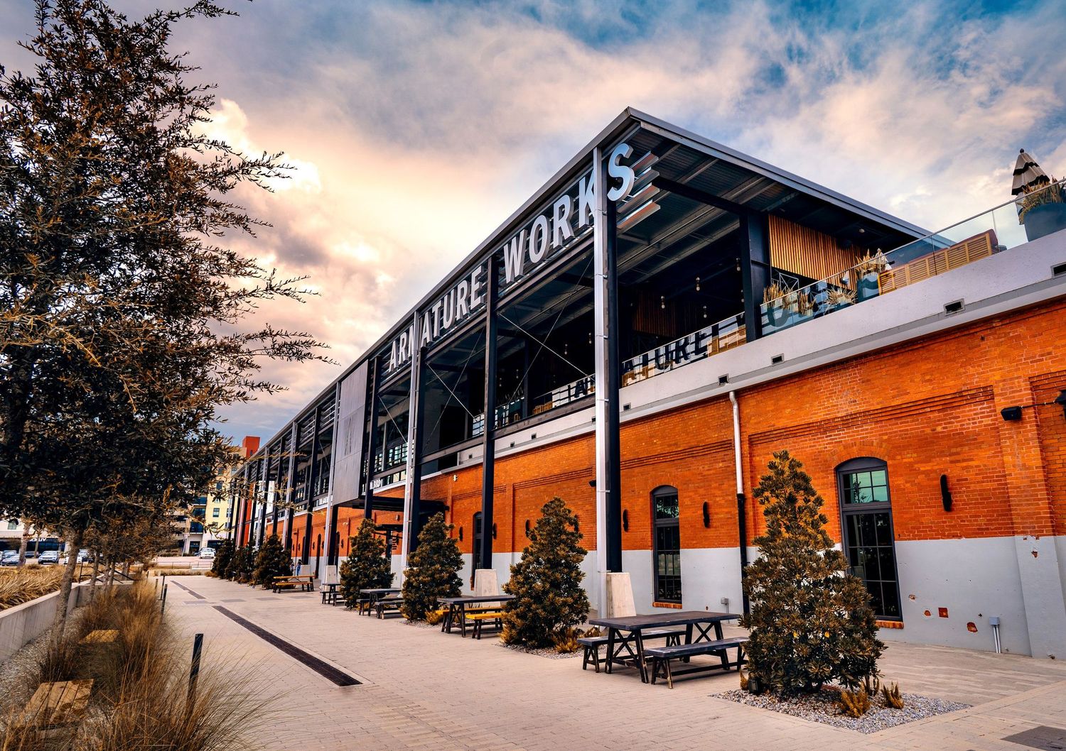 Red brick industrial building with Works signage, steel canopy, potted evergreens and picnic tables along a paved walkway at sunset.
