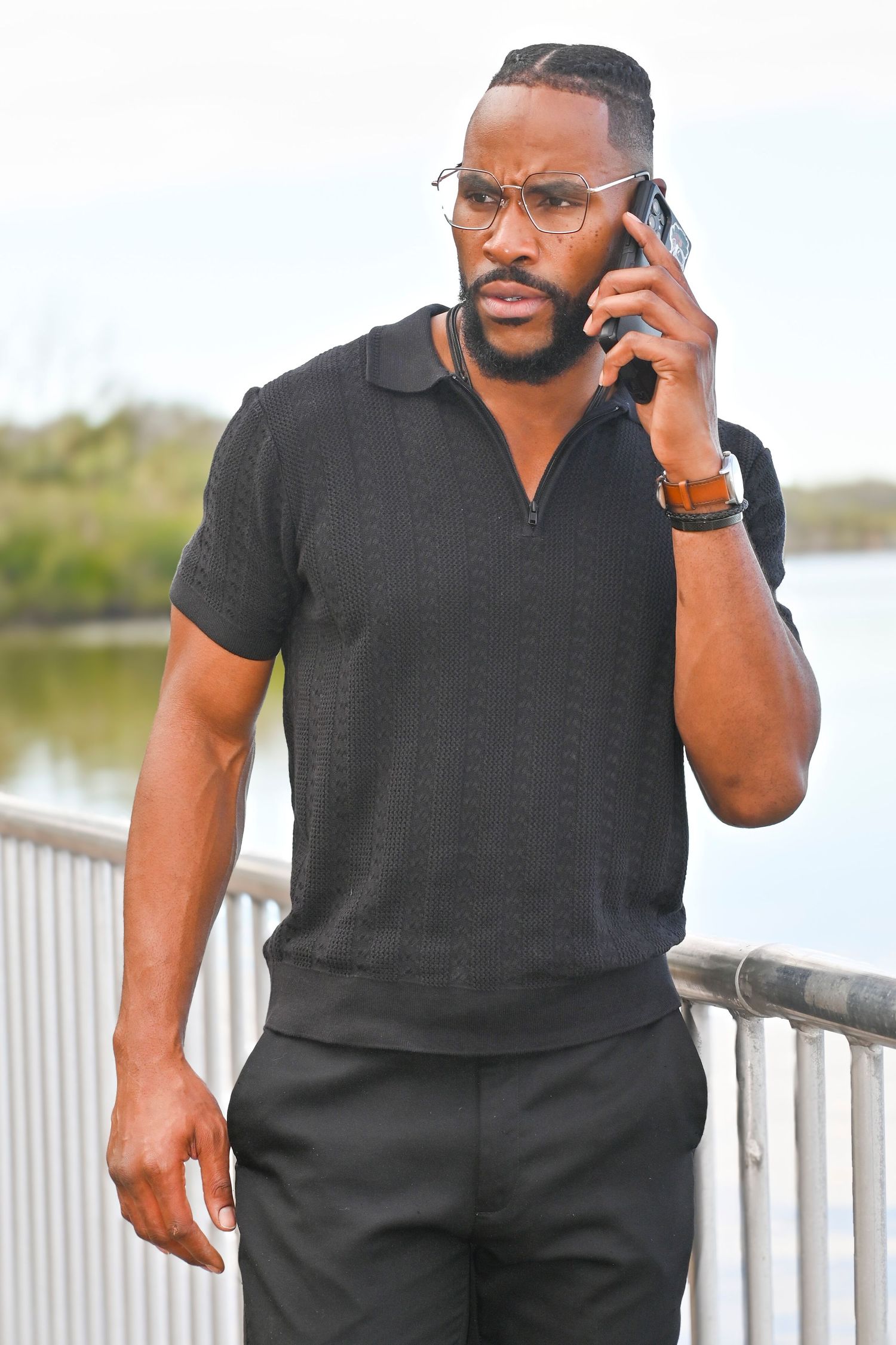 Bearded man in glasses and a black knit polo talks on a smartphone while standing beside a metal railing with a calm waterway behind him.