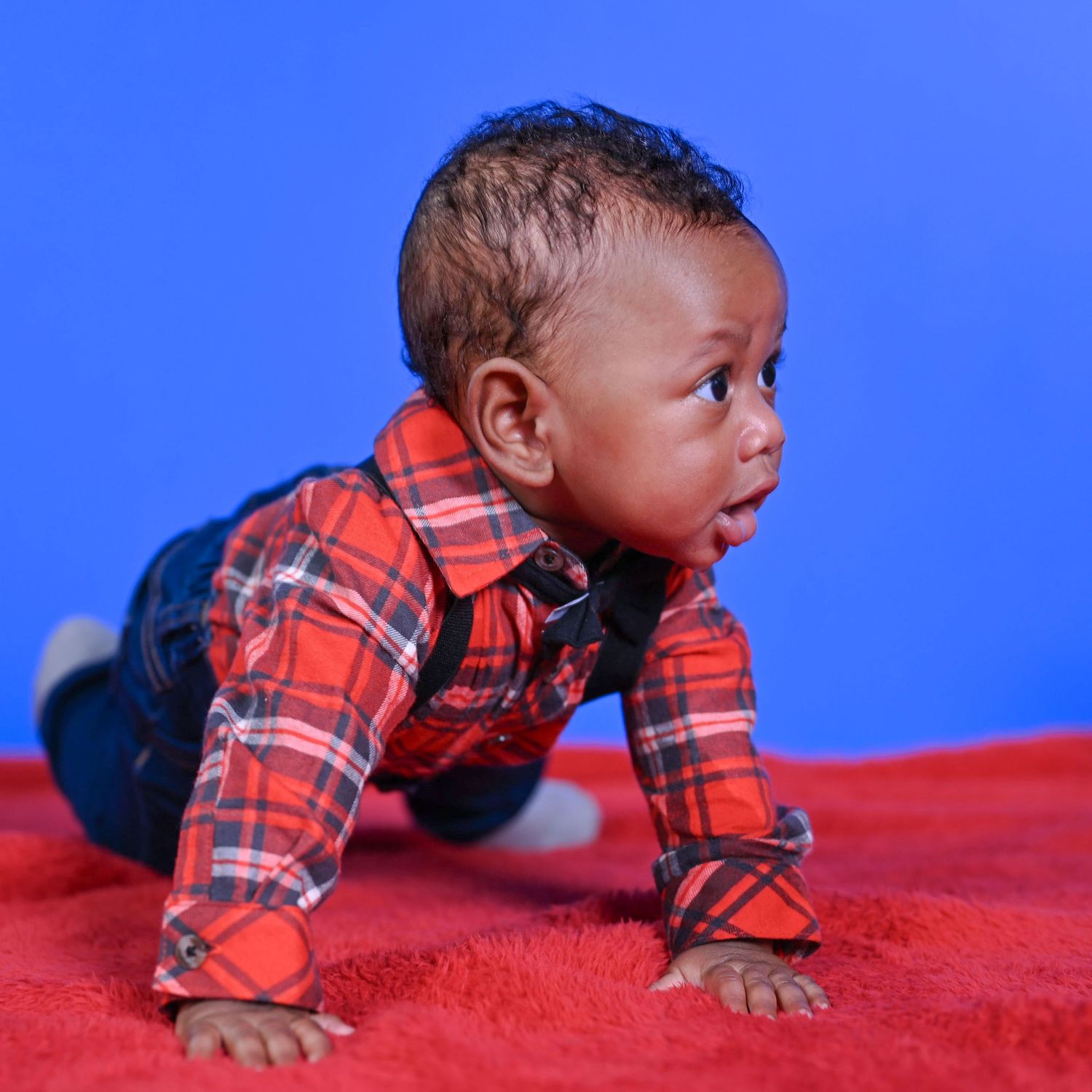 Baby in a red plaid shirt and denim overalls crawling on a red blanket against a bright blue backdrop.
