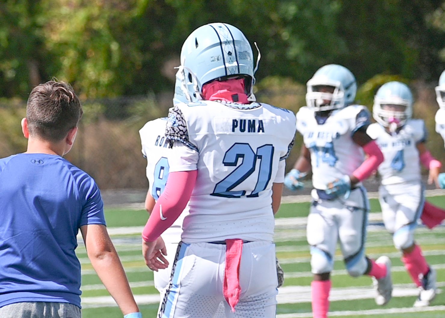 Football player in white jersey with pink accessories stands on sideline during game.