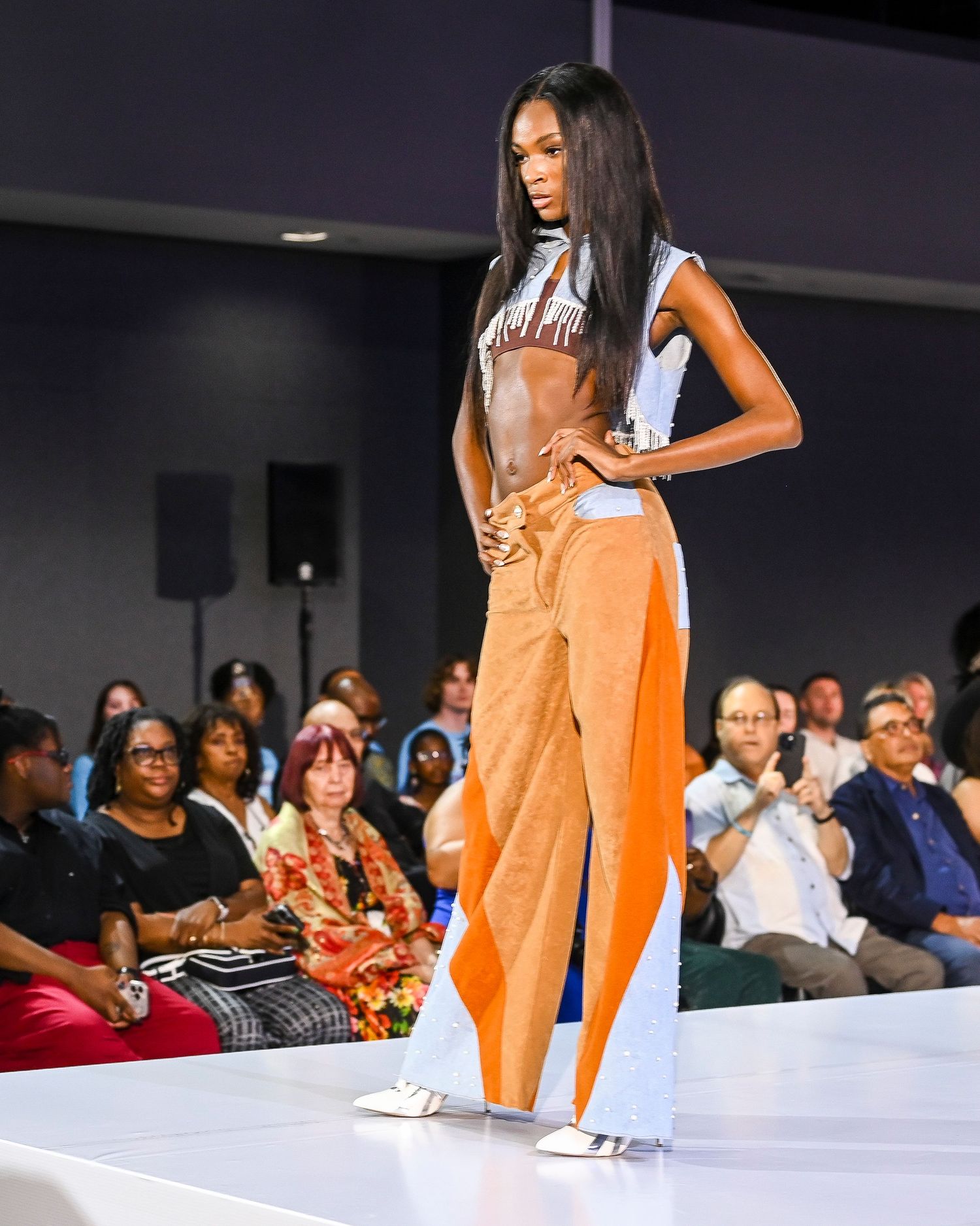 Models showcase orange fashion designs on white runway with audience.