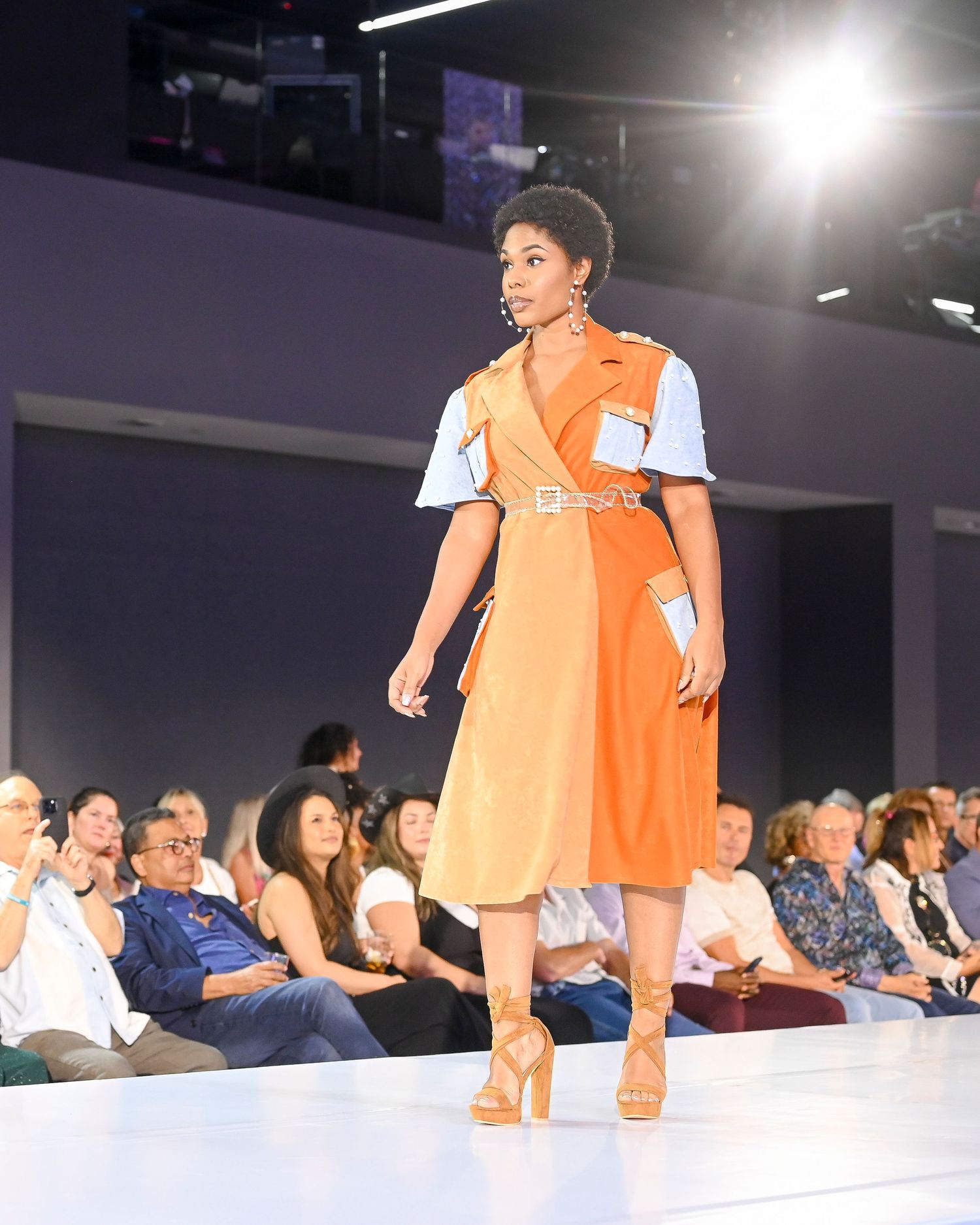 Fashion runway show with orange outfit and audience in background.