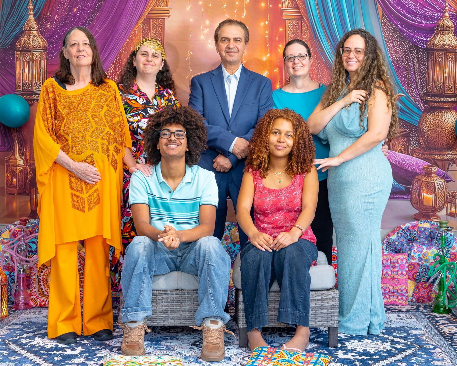 Seven people pose together in colorful, vibrant attire against a decorative backdrop with lanterns, drapes, and ornate rugs.