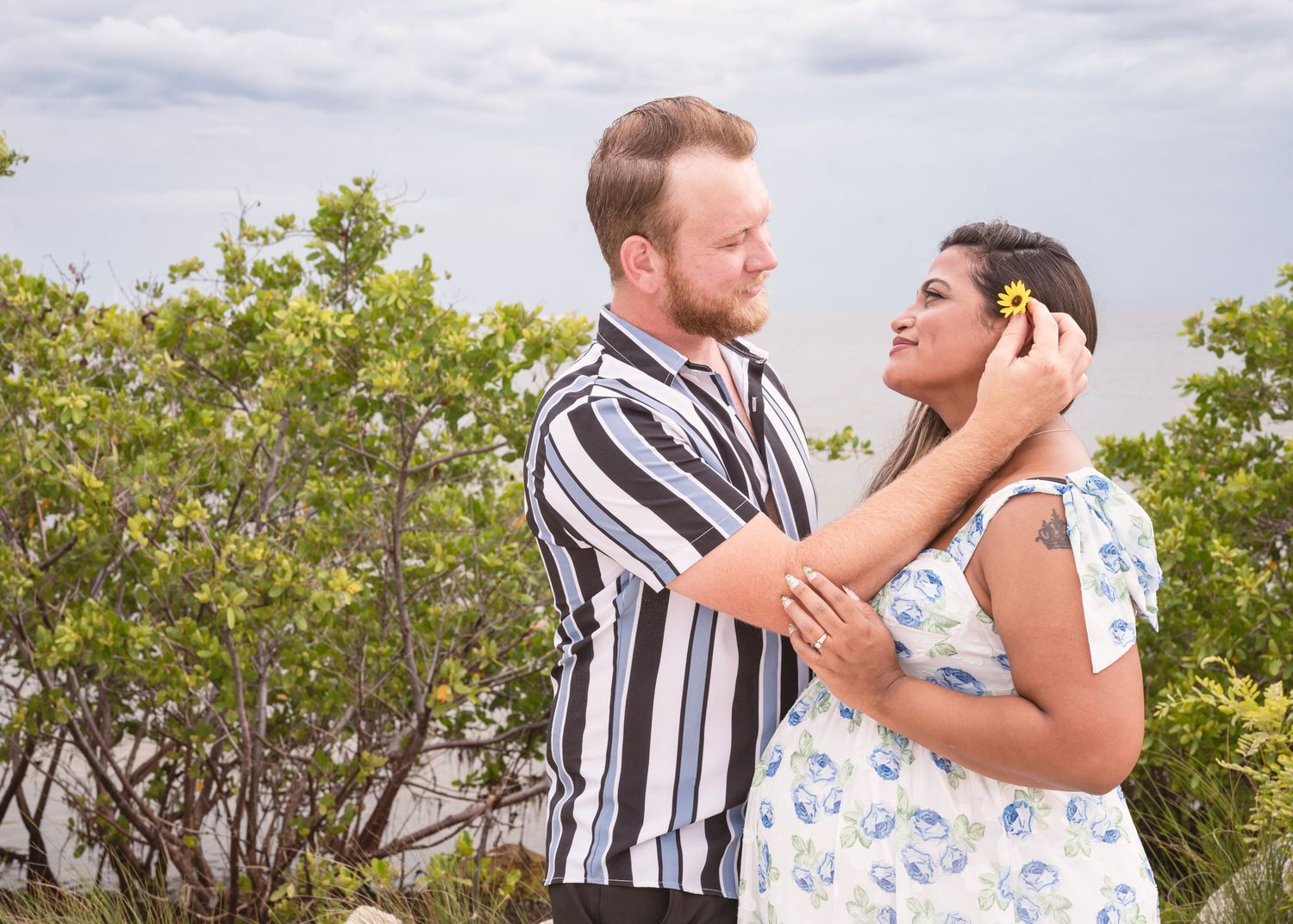 A couple shares a romantic moment outdoors, surrounded by lush green trees under a cloudy sky.