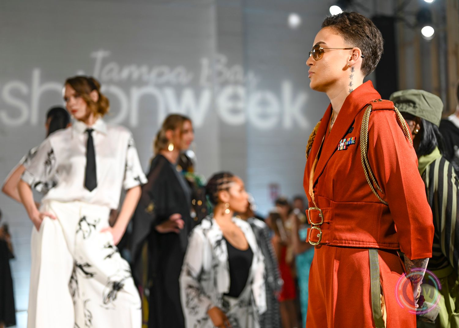 Models in stylish outfits, including a rust-orange jacket and a white printed dress, pose at Tampa Bay Fashion Showweek.