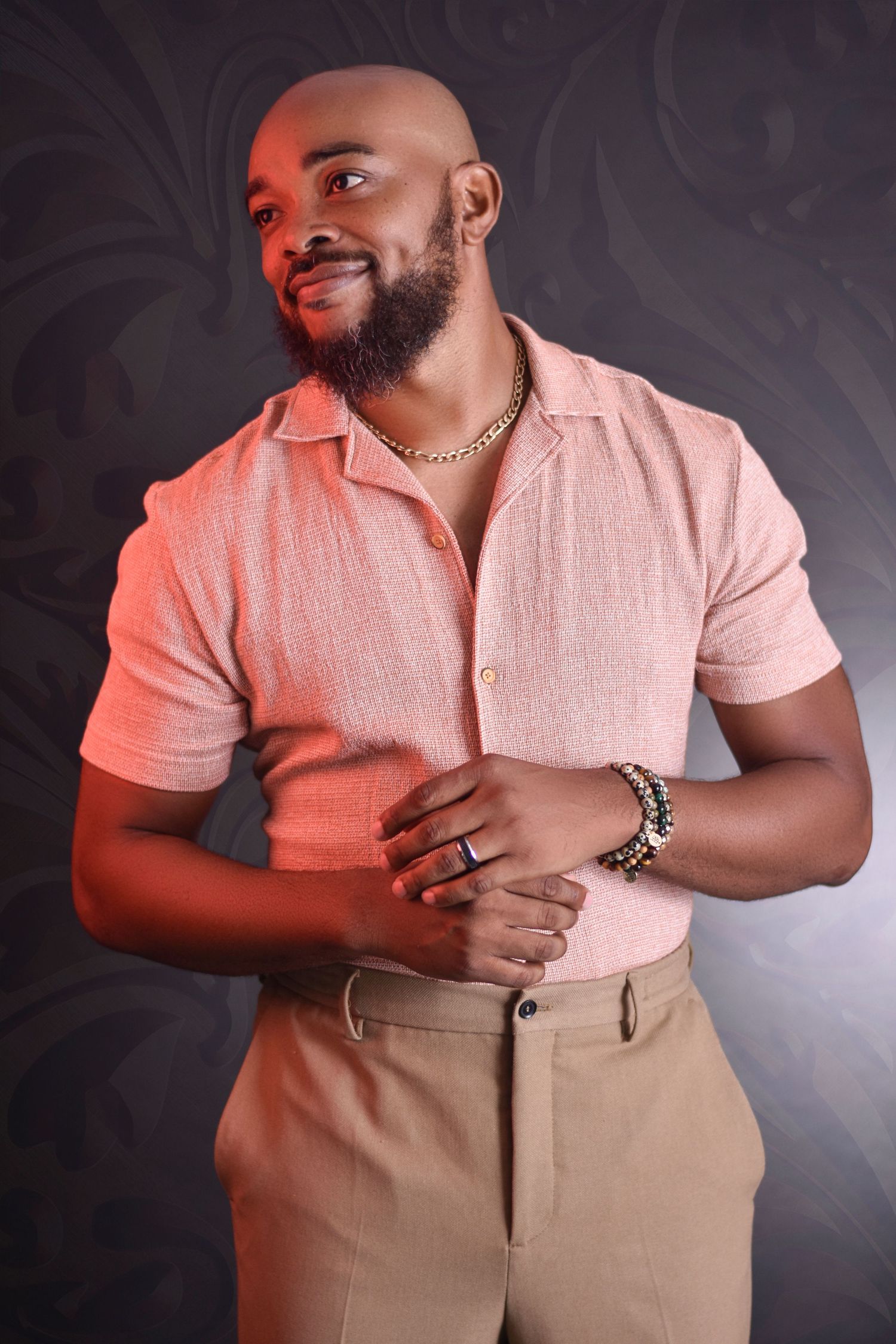 Studio portrait in pink polo shirt and khaki pants against dark patterned background with dramatic lighting.