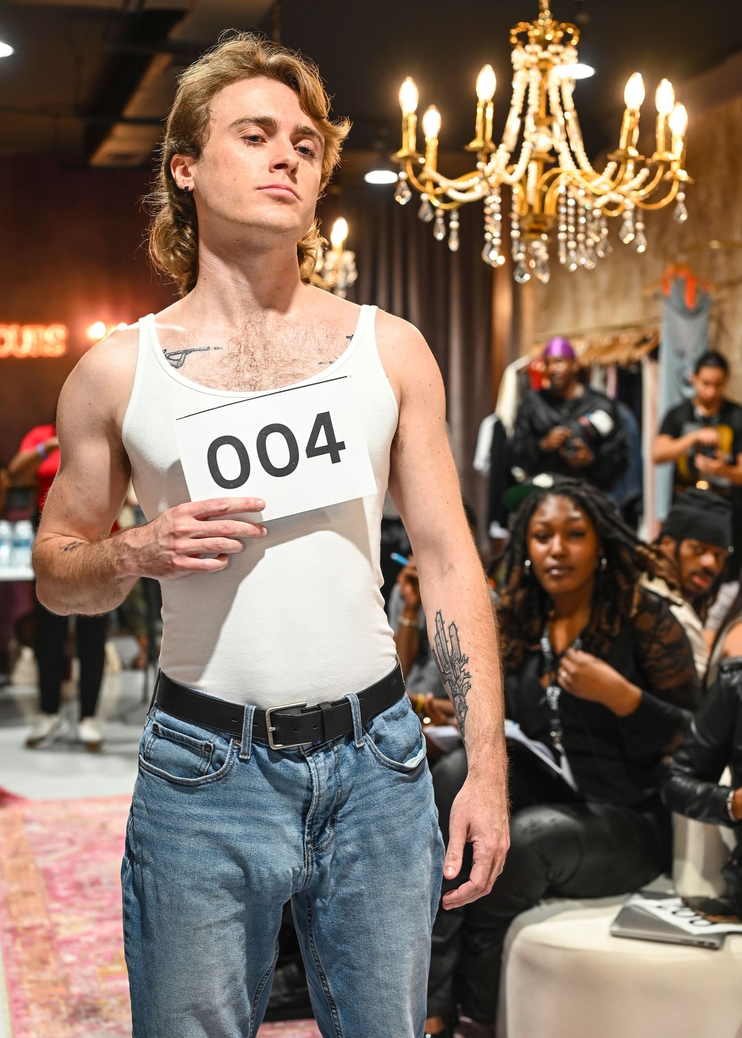Models walking the runway in classic white tank tops and blue jeans under crystal chandeliers.