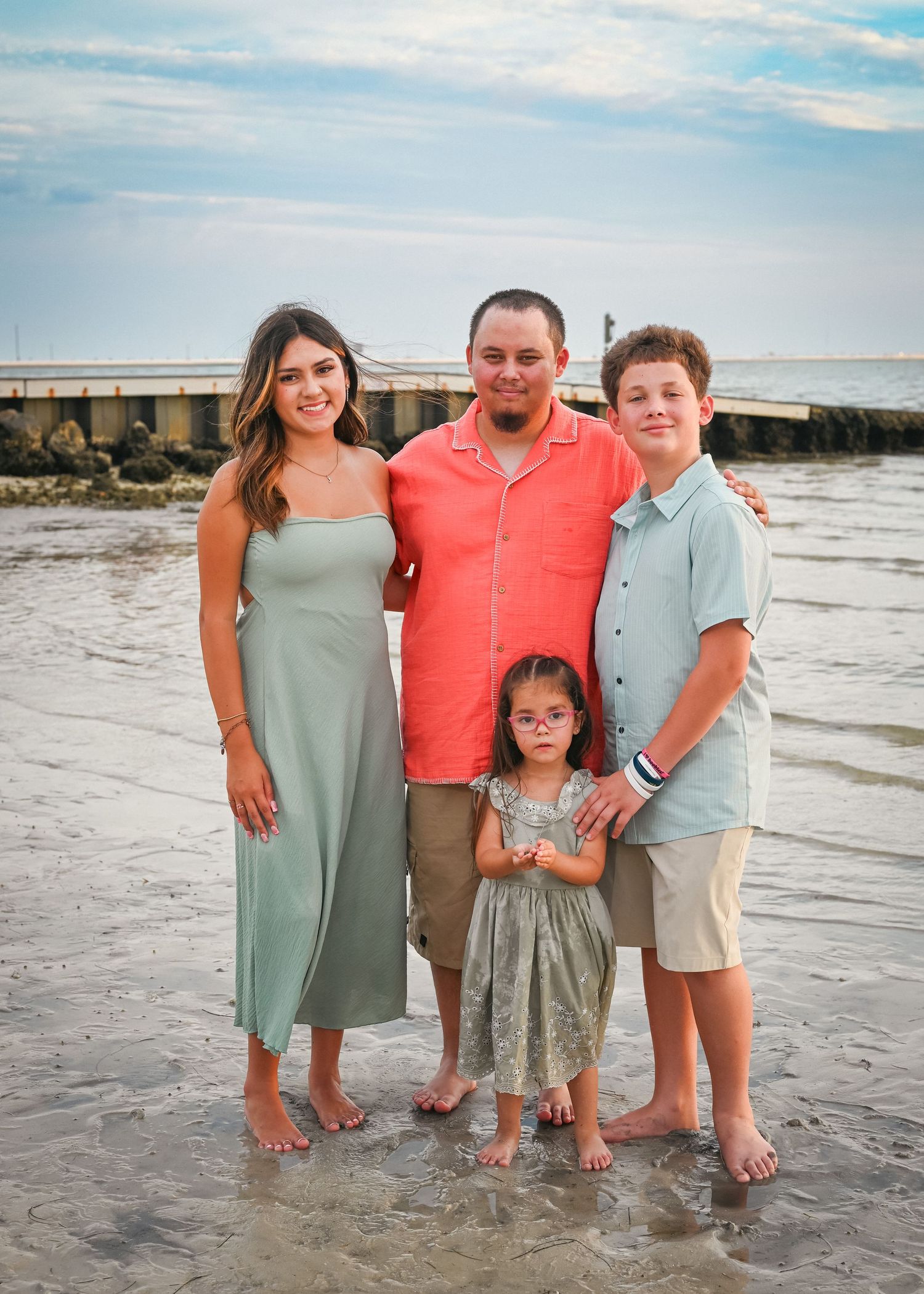 Family of four posing on a beach at sunset, with a pier in the background, wearing casual summer outfits.