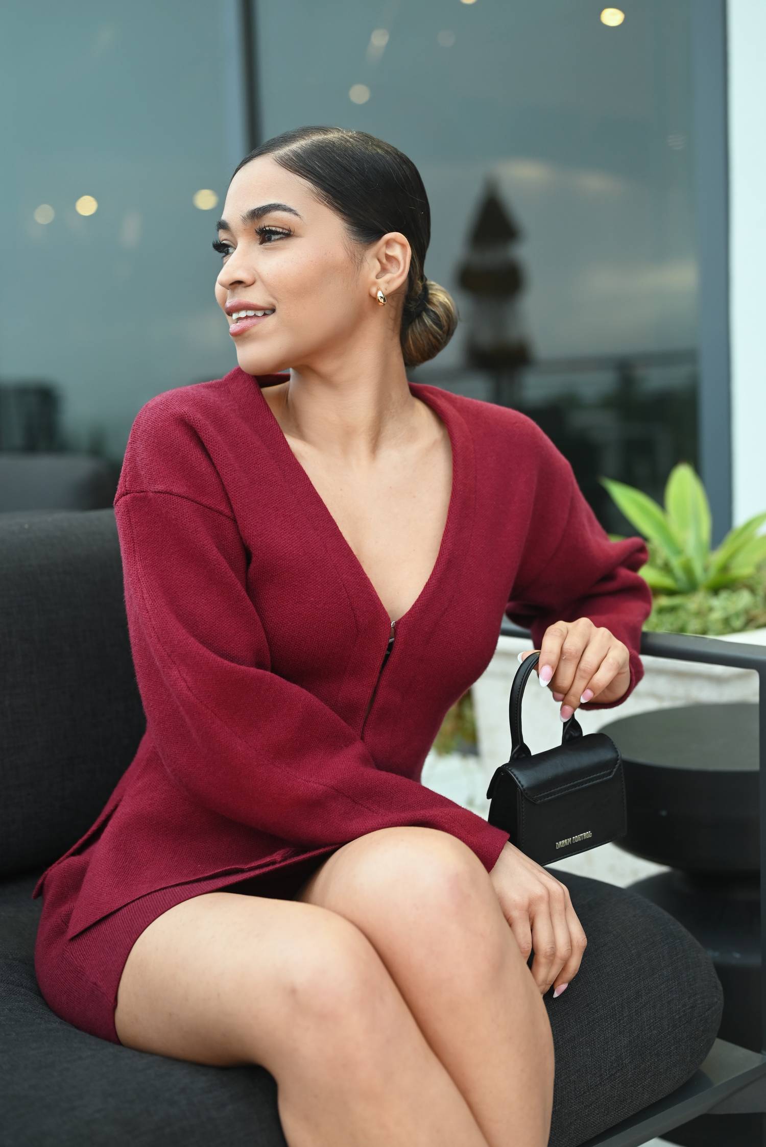 Professional model in burgundy wrap dress poses elegantly on dark couch with modern decor in background.