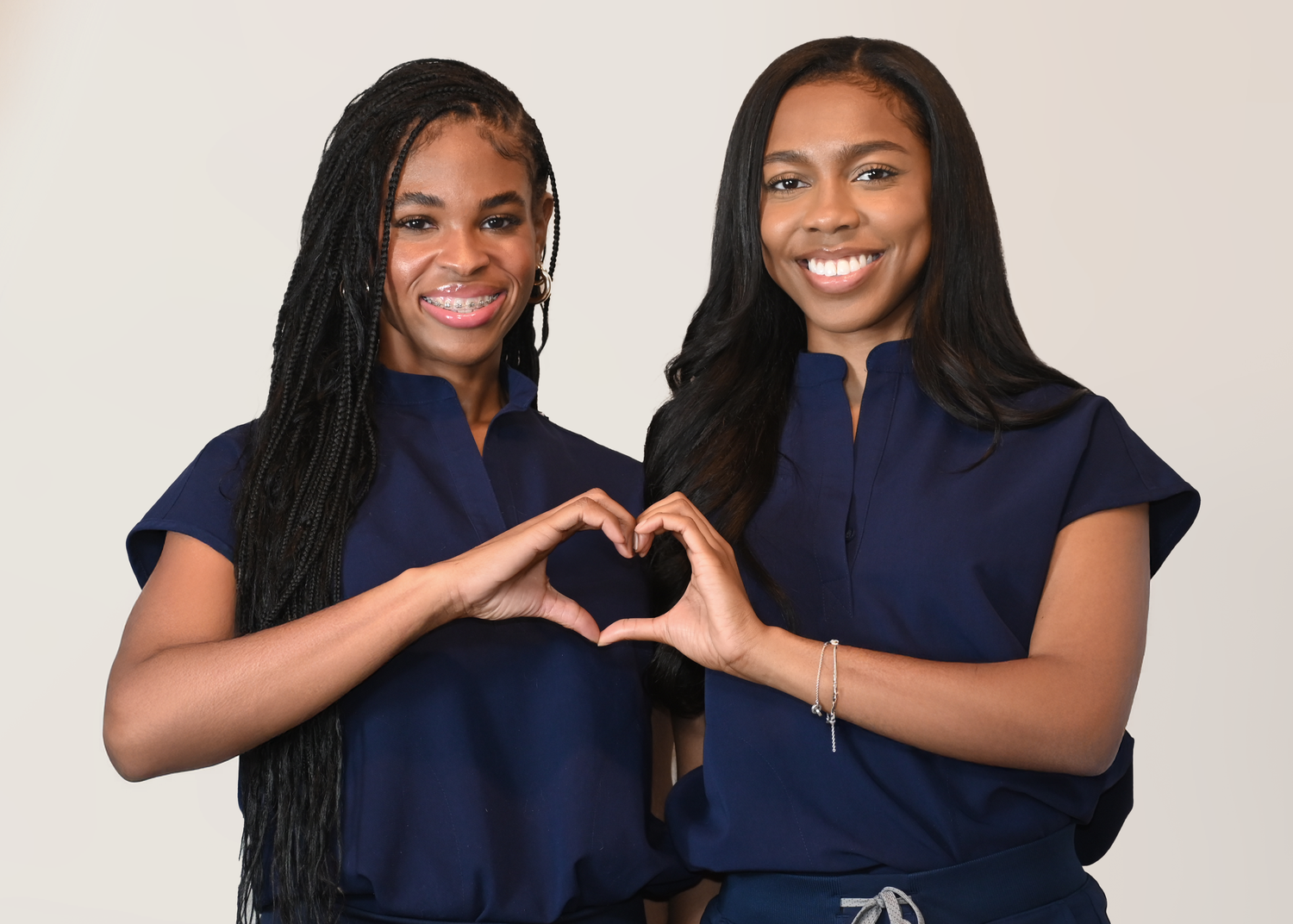 Two people in navy scrubs make heart shapes with their hands while smiling at the camera.