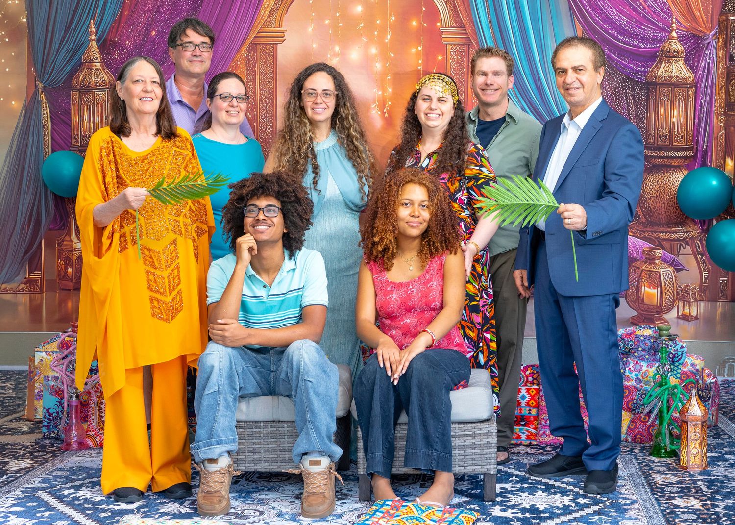 Group of nine people posing together in front of a colorful Moroccan-themed backdrop, some holding palm leaves.