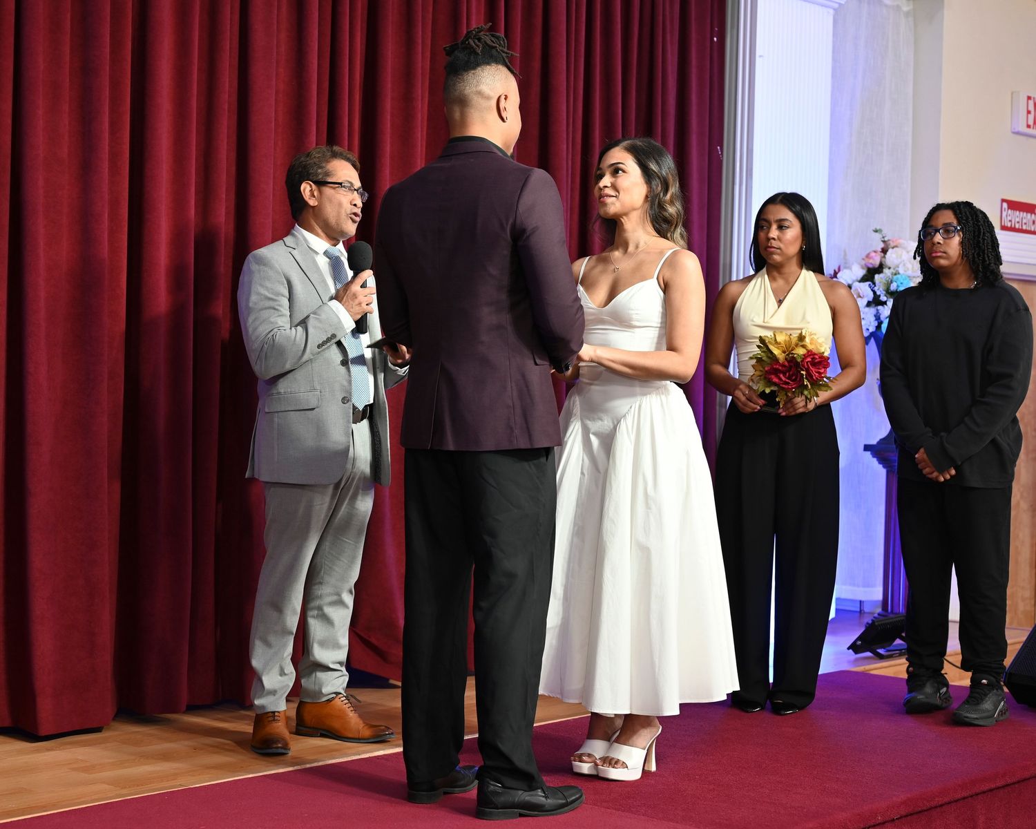 Wedding ceremony scene with formal attire against red curtains and white walls.