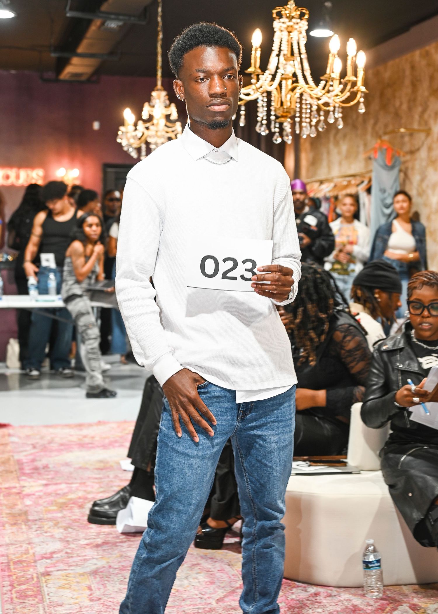 Models walk the runway in white tops and jeans under elegant chandeliers at a fashion show.