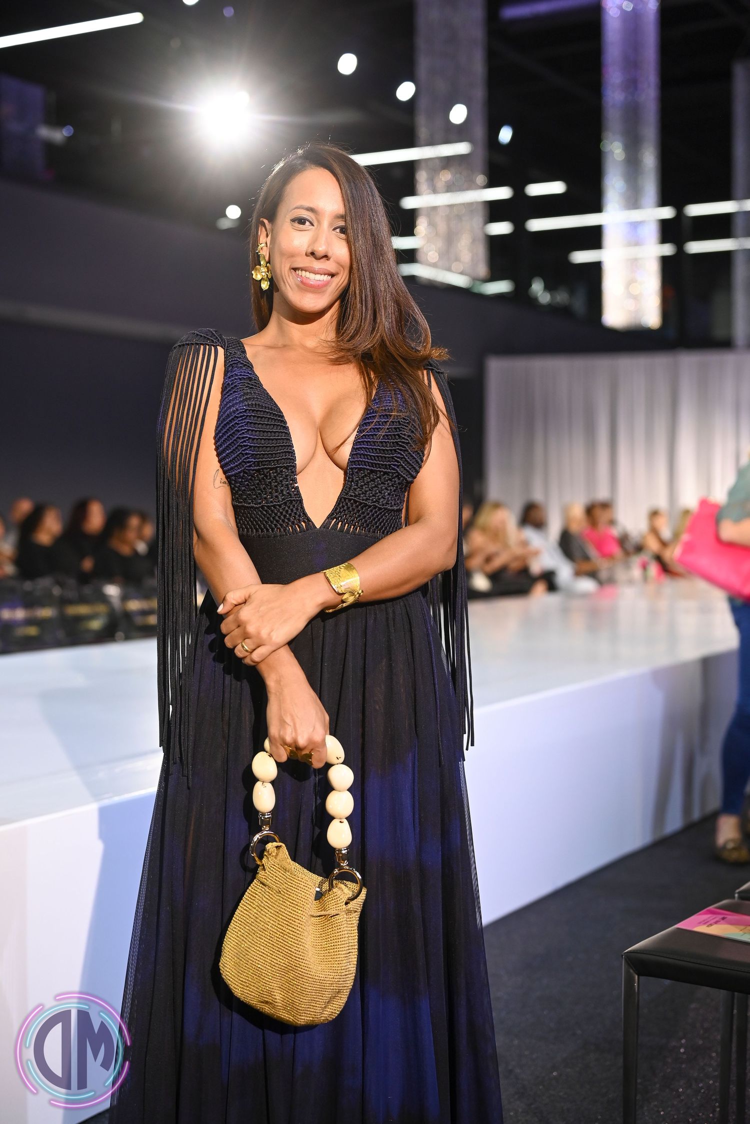 Navy blue dress with fringe details and gold handbag accessory shown on fashion runway with studio lighting.