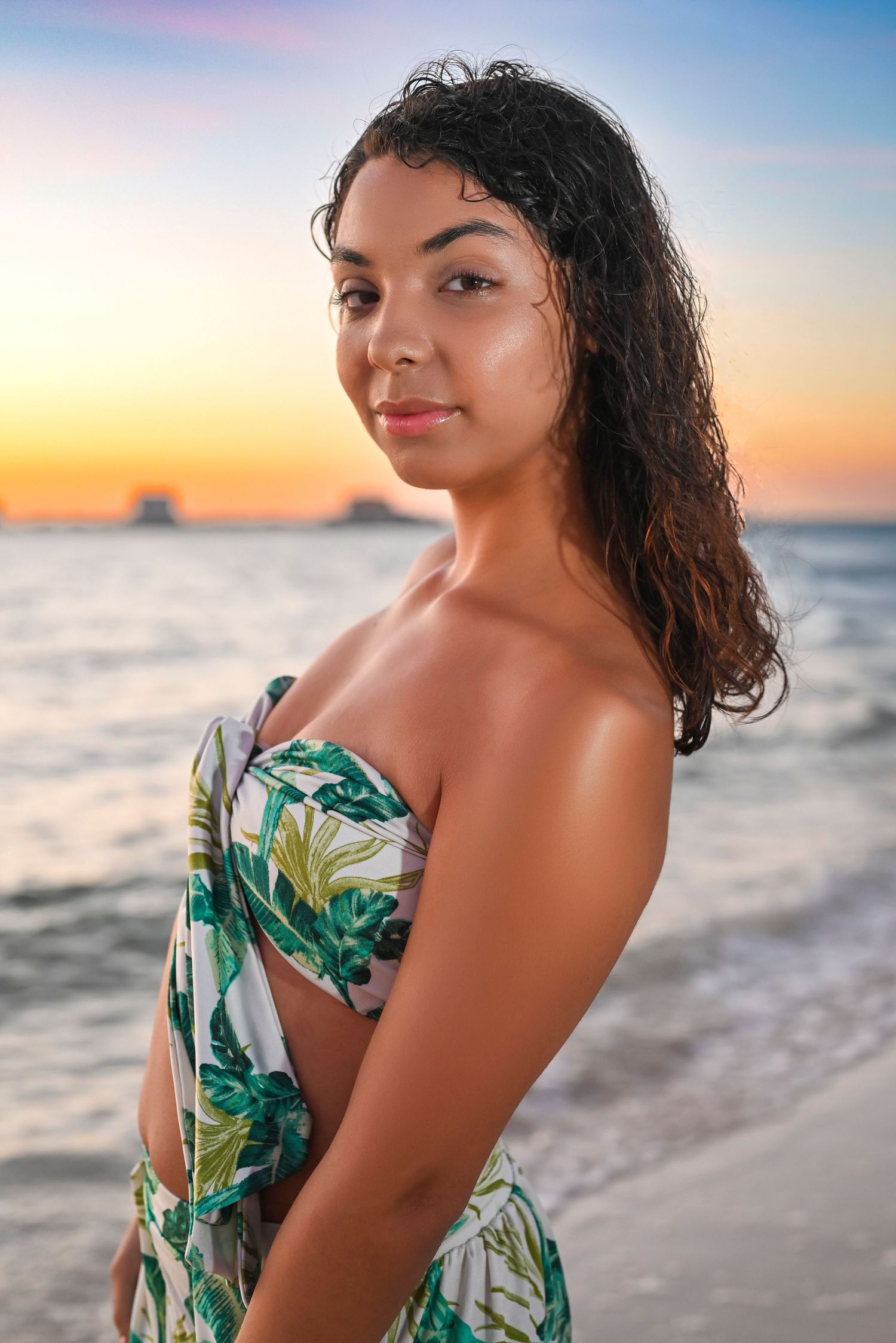 Woman in tropical print dress posing at beach during sunset.