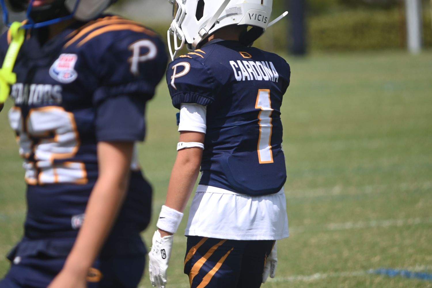 Youth football player wearing white helmet and navy number 1 jersey with Cardona on the back stands on a grassy field beside a teammate.