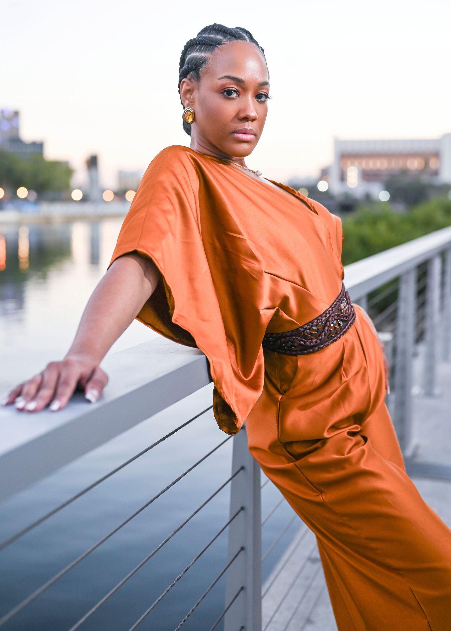 Woman in an orange satin dress with braided updo leaning on a metal railing beside a waterfront at dusk.