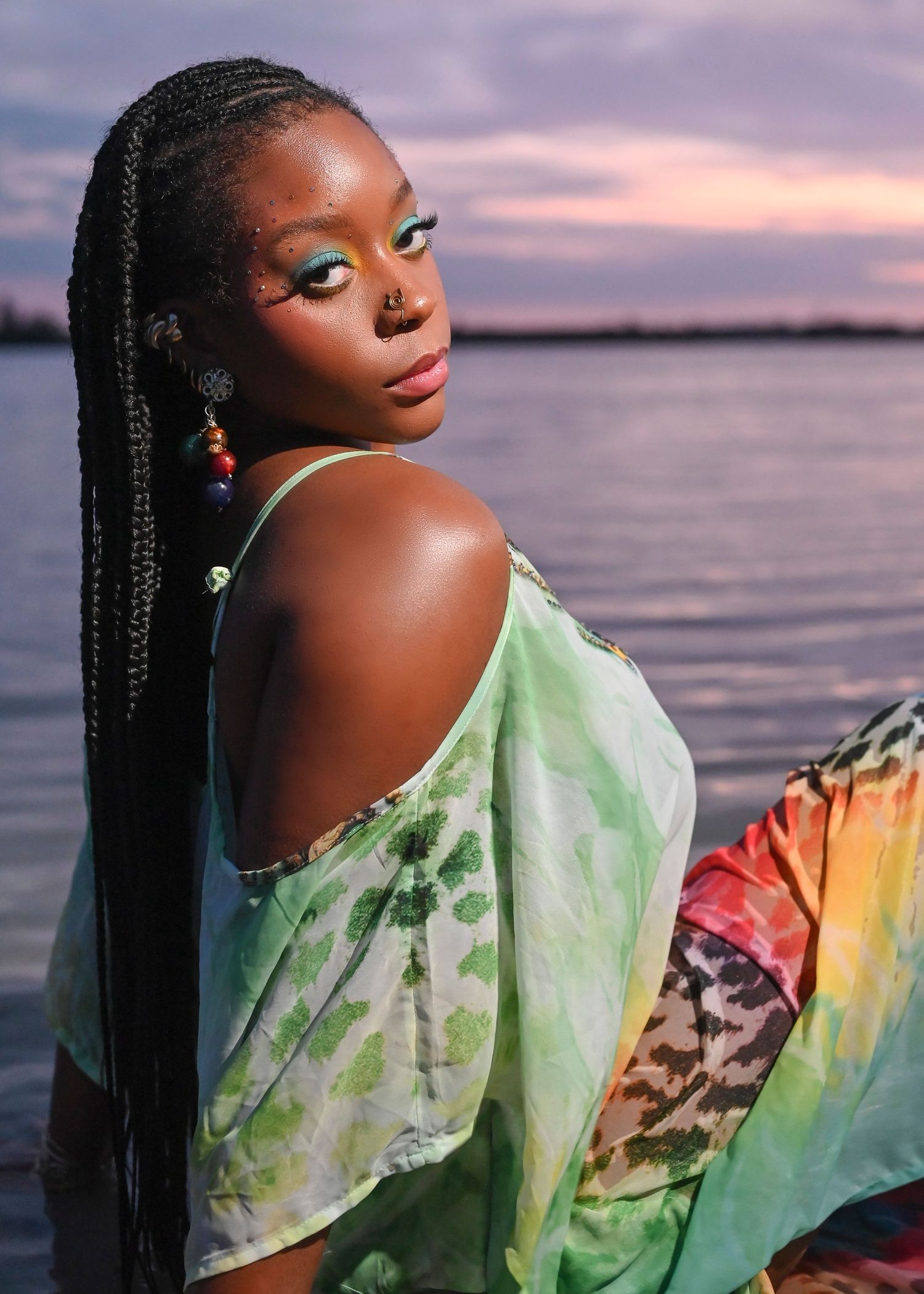 Woman with long braids and colorful eye makeup wearing a tie-dye green dress poses near water at sunset.