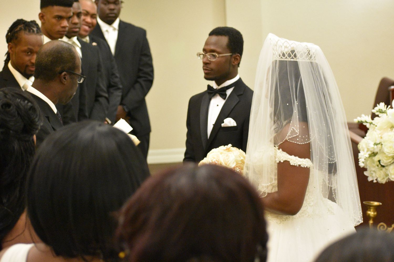 A couple exchanges vows at a formal wedding ceremony, bride in white veil and groom in tuxedo, surrounded by groomsmen.