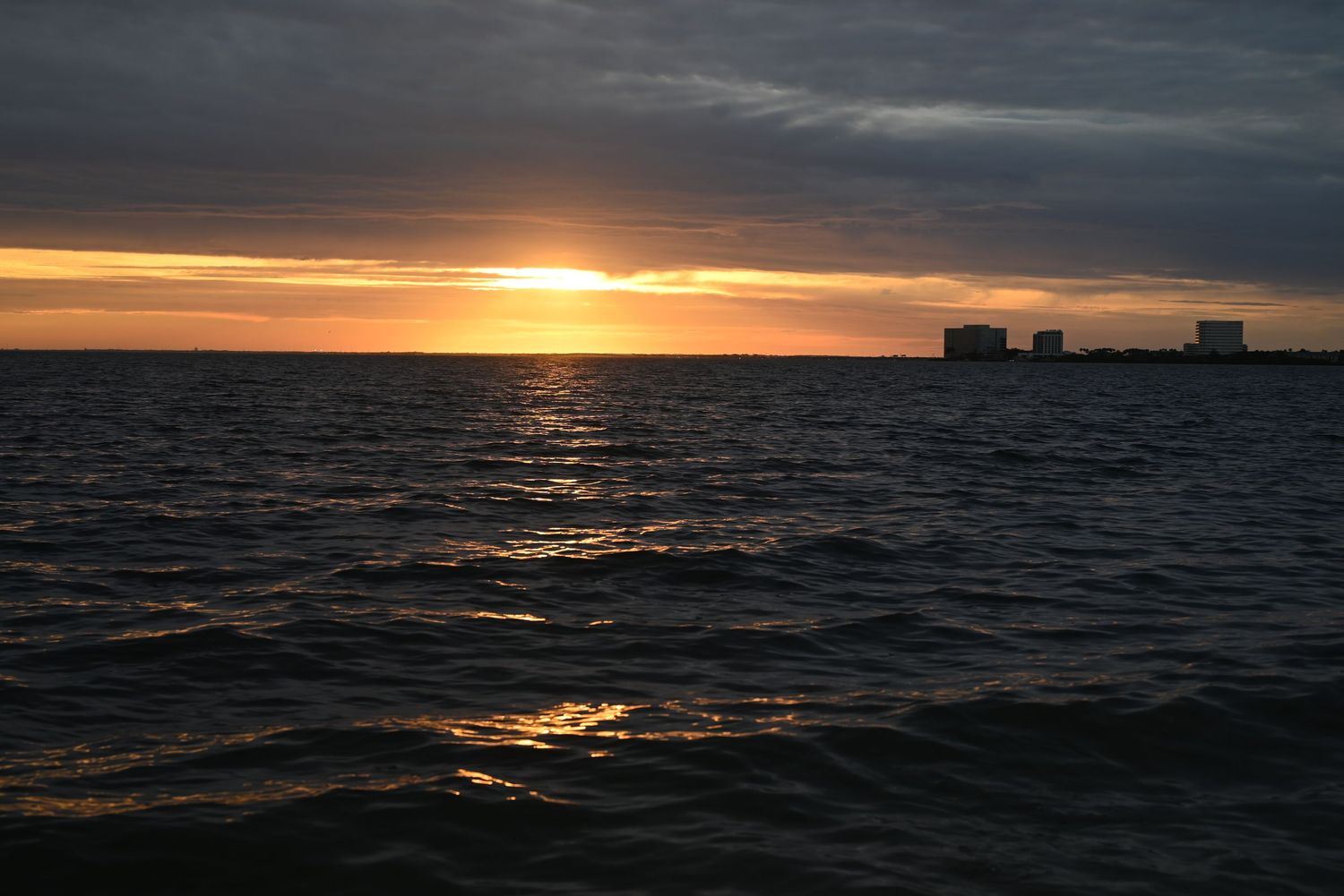 Stunning ocean sunset with golden sun on horizon, dark choppy waters, and distant buildings silhouetted against colorful sky.