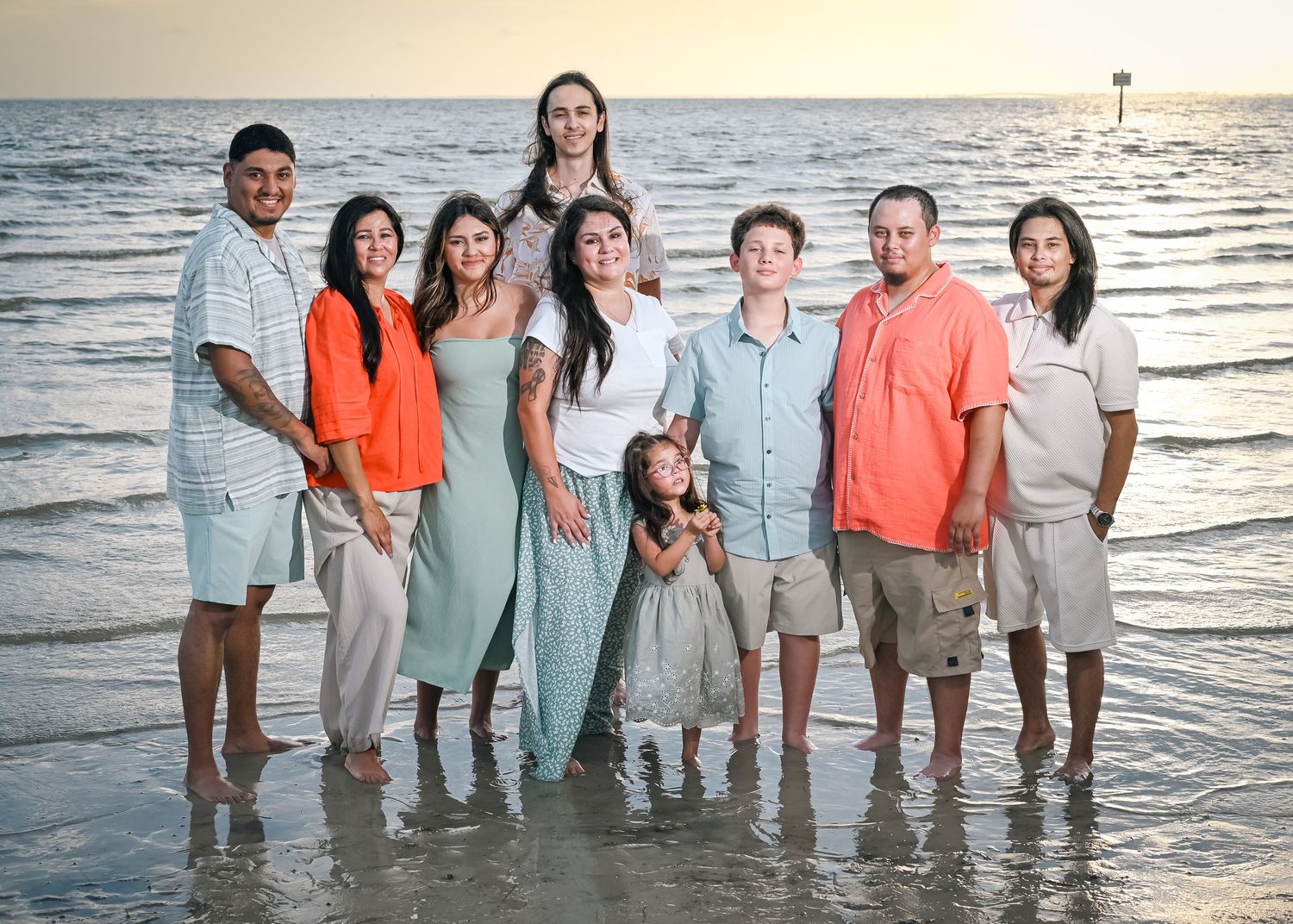 Family of nine posing together on a sandy beach at sunset with calm ocean waves around their feet.
