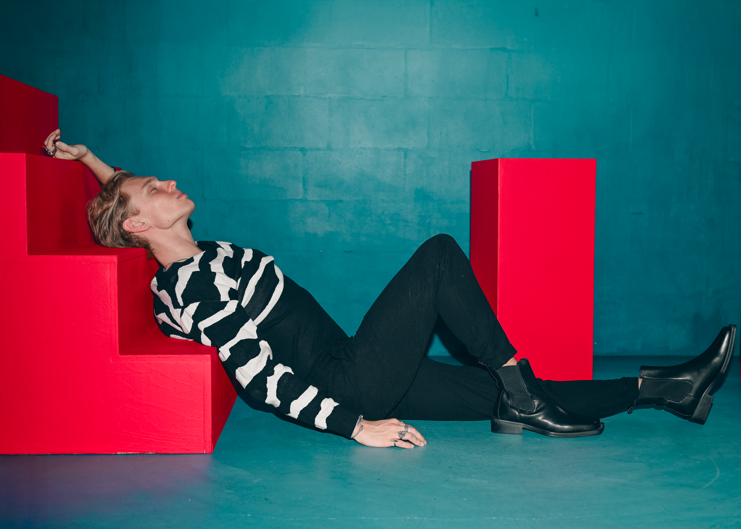 Person in a black and white striped sweater, black pants and boots reclining against bold red geometric blocks on a teal studio backdrop.