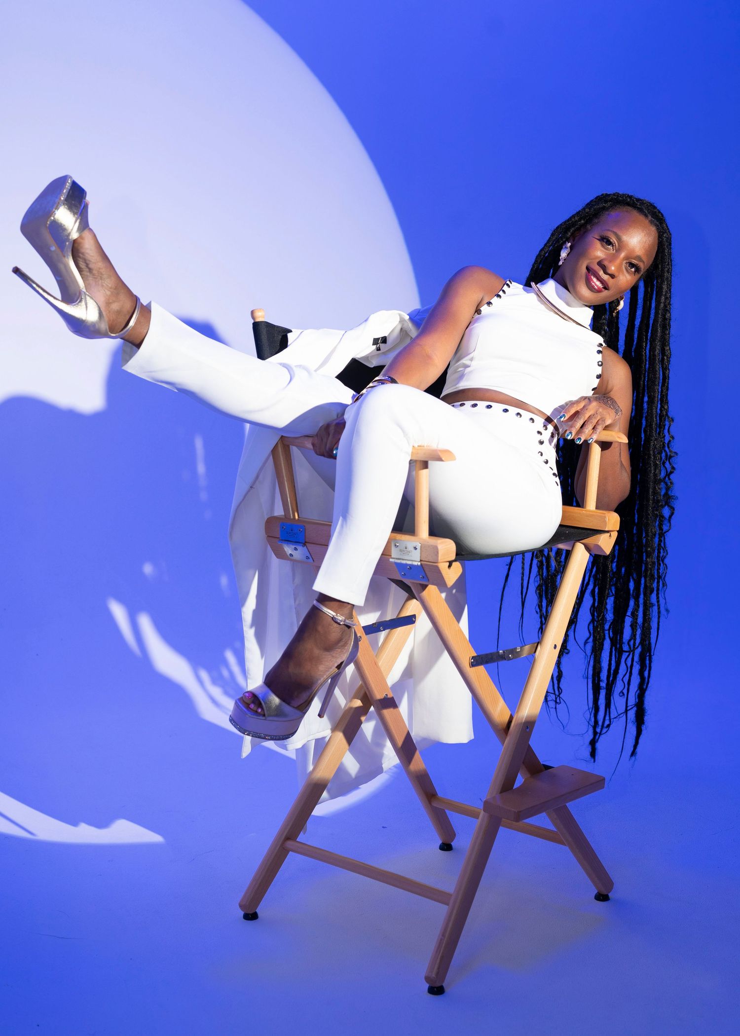 Fashion model in white outfit strikes playful pose on wooden chair with blue background.