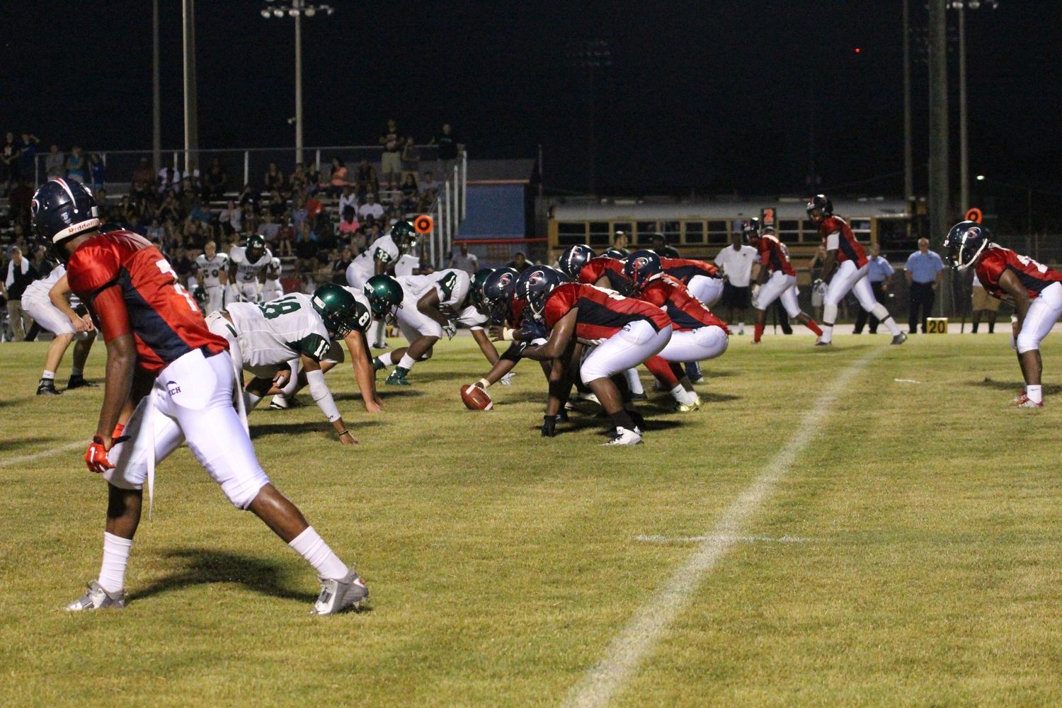 High school football players in red and white and green uniforms line up at the line of scrimmage during a night game under stadium lights.