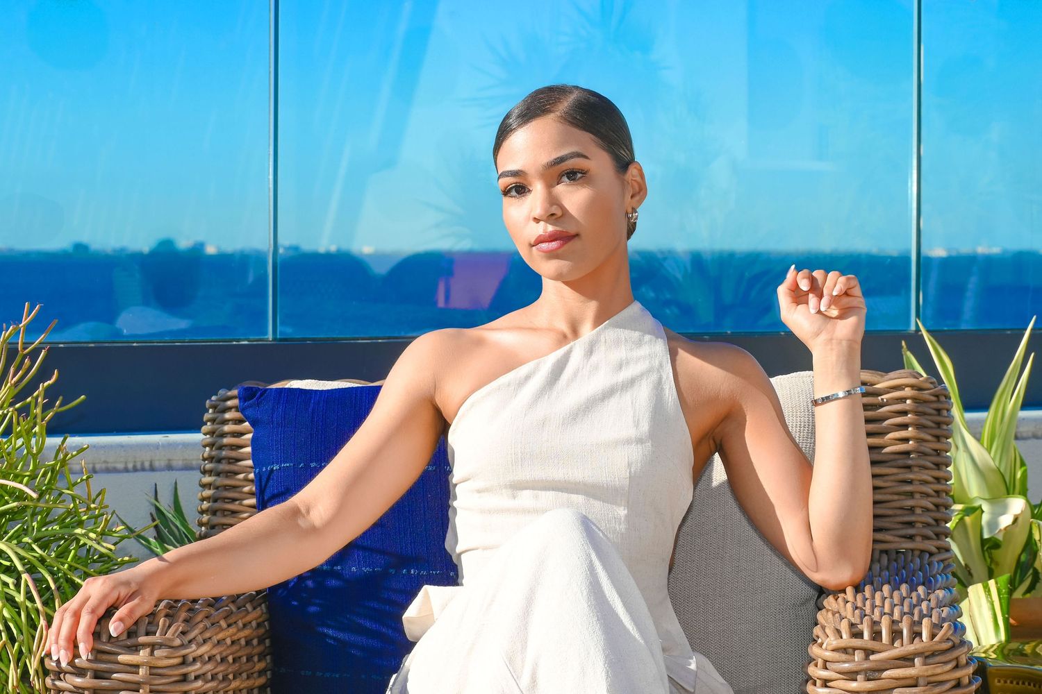 Elegant woman in white dress seated on blue cushioned patio furniture against blue glass backdrop.