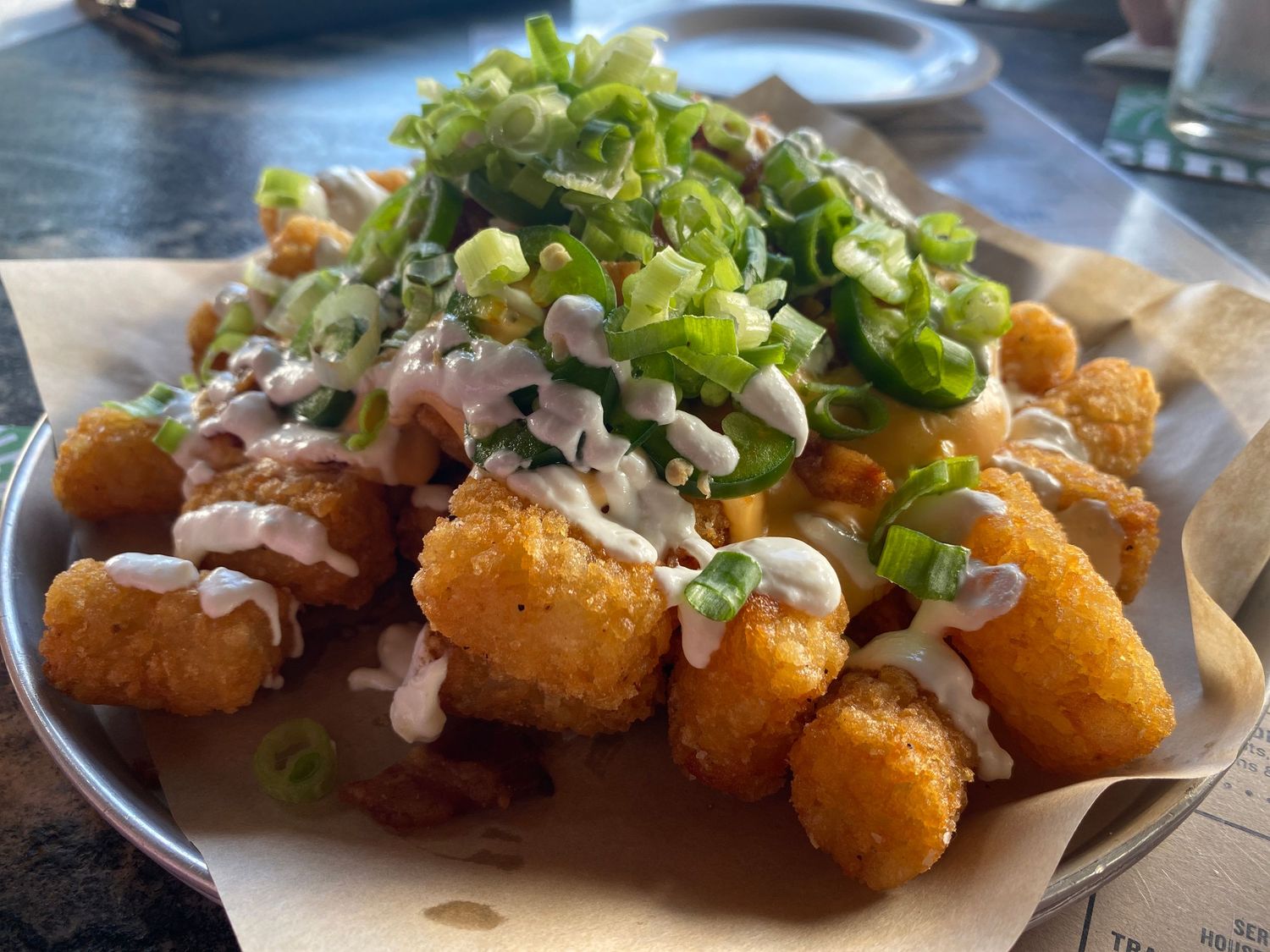 Crispy tater tots topped with green onions and sauce on serving paper.