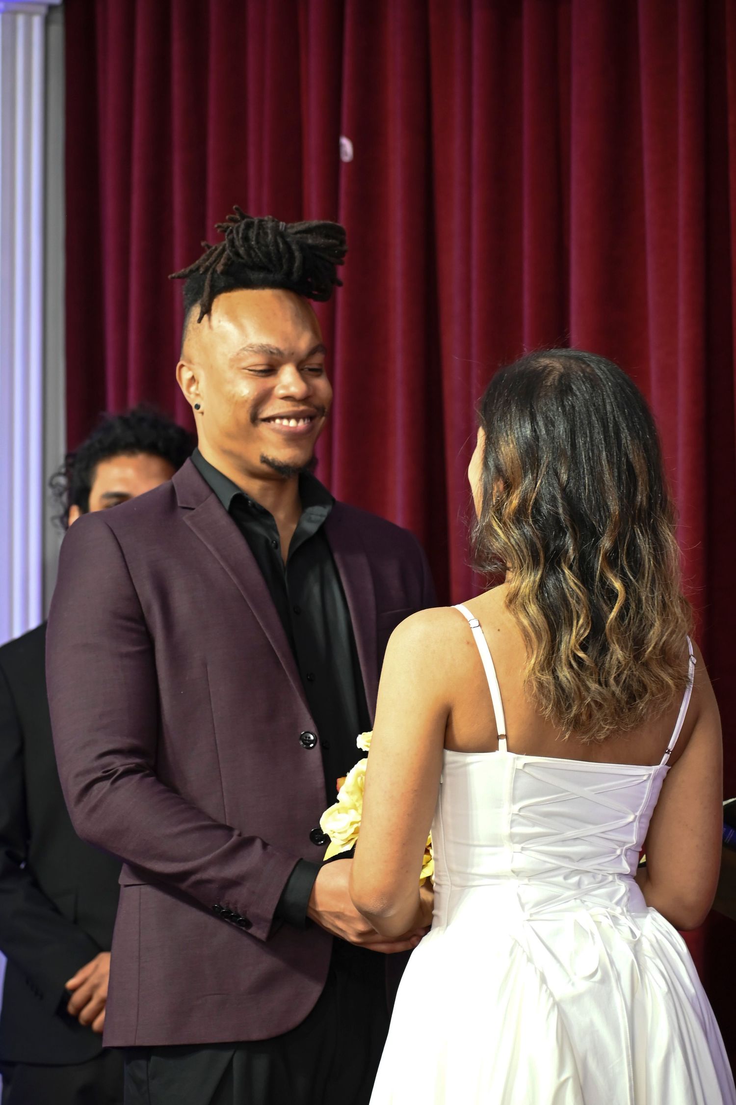 Wedding ceremony scene with couple in formal attire against red curtain backdrop.