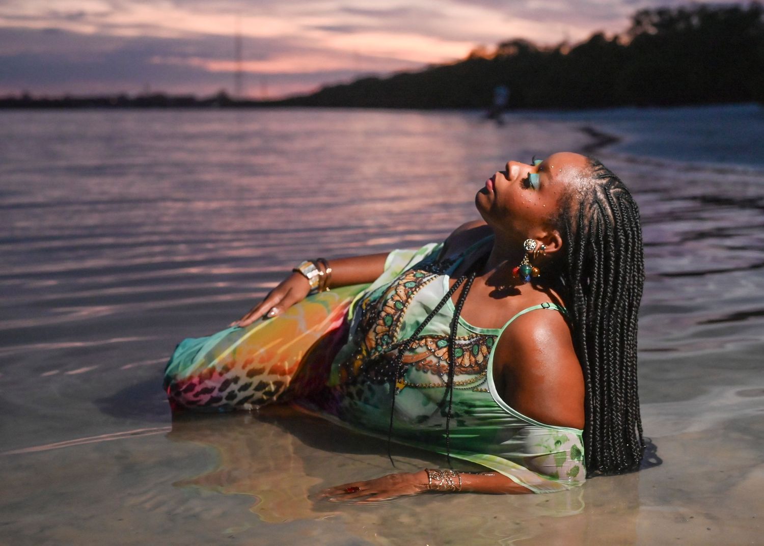 Woman with braids reclines in shallow water at sunset, wearing a colorful dress, head tilted back peacefully.