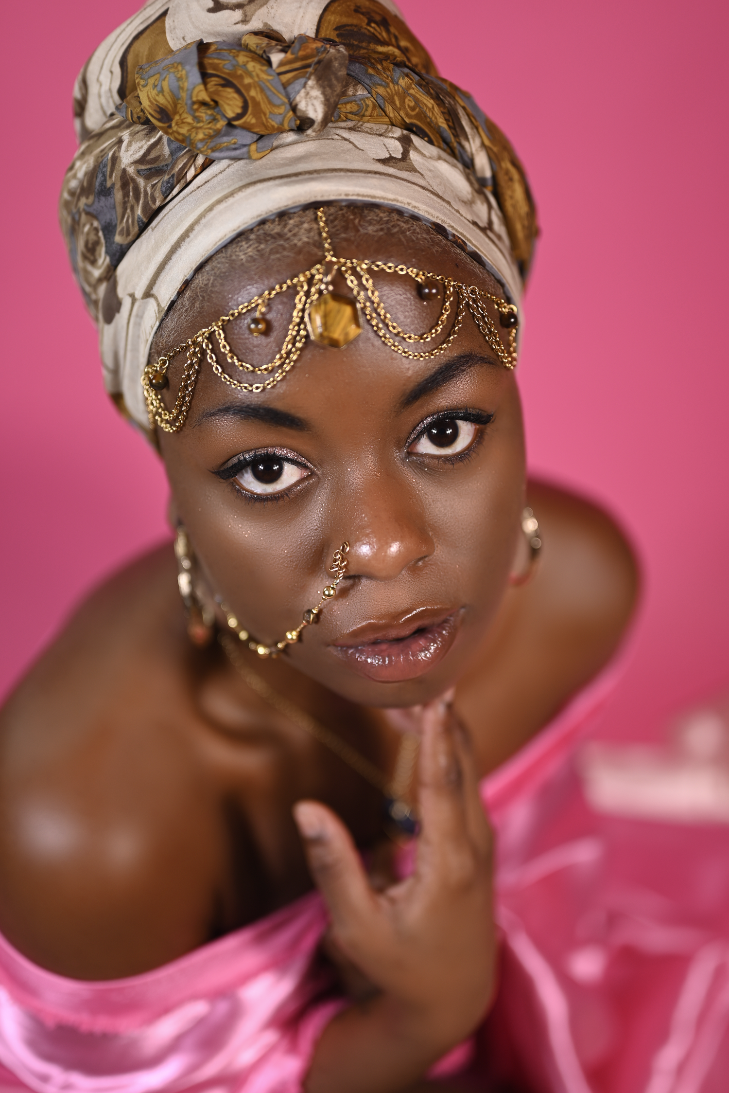 Artistic portrait against pink backdrop featuring gold accessories and head wrap.