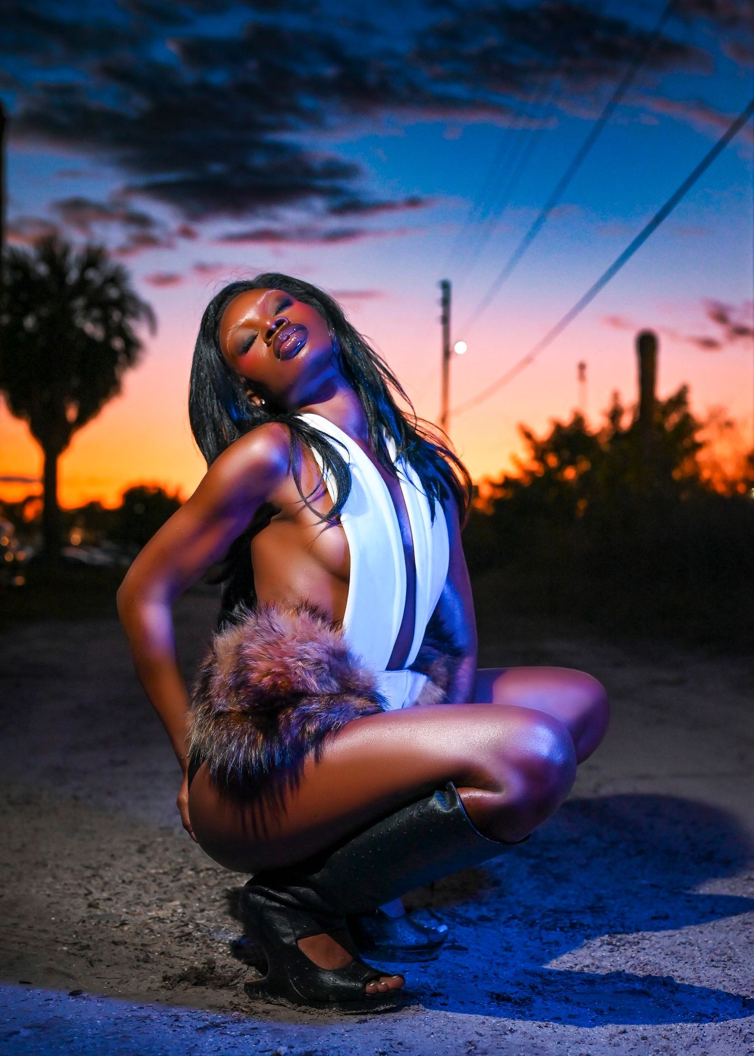 A woman poses confidently at sunset in a white bodysuit and fur accessory on a dramatic blue and orange twilight street.