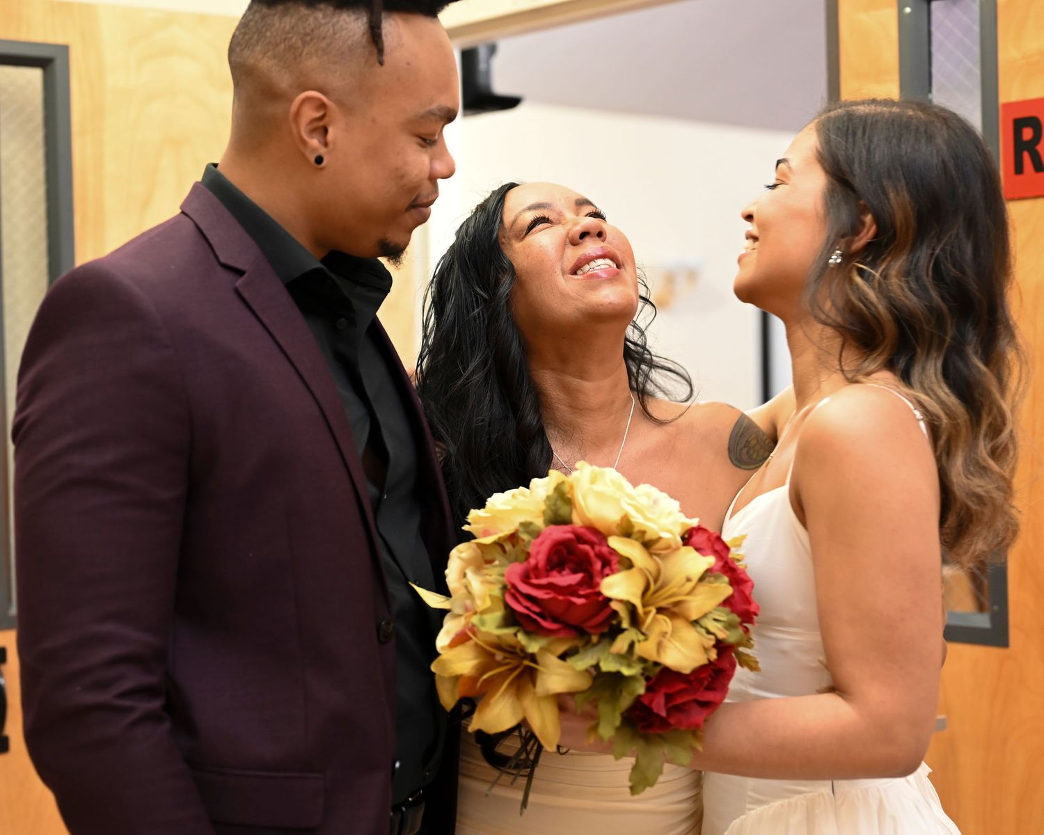A joyful wedding moment with the couple and bridesmaid holding a bouquet of red and gold flowers.