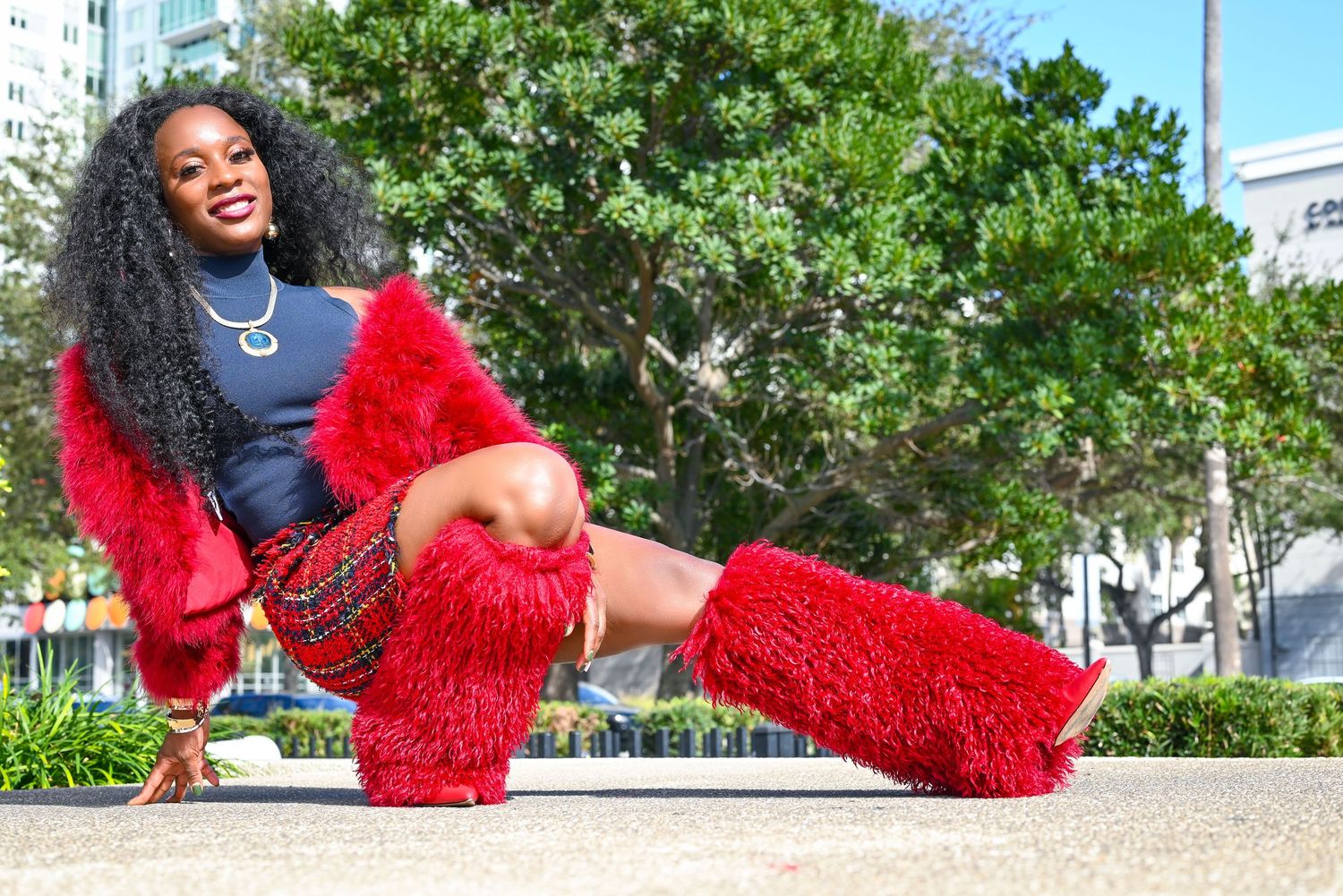 Woman with long curly hair poses seated outdoors in red plaid skirt, fuzzy red leg warmers and fur sleeves, with green trees behind her.