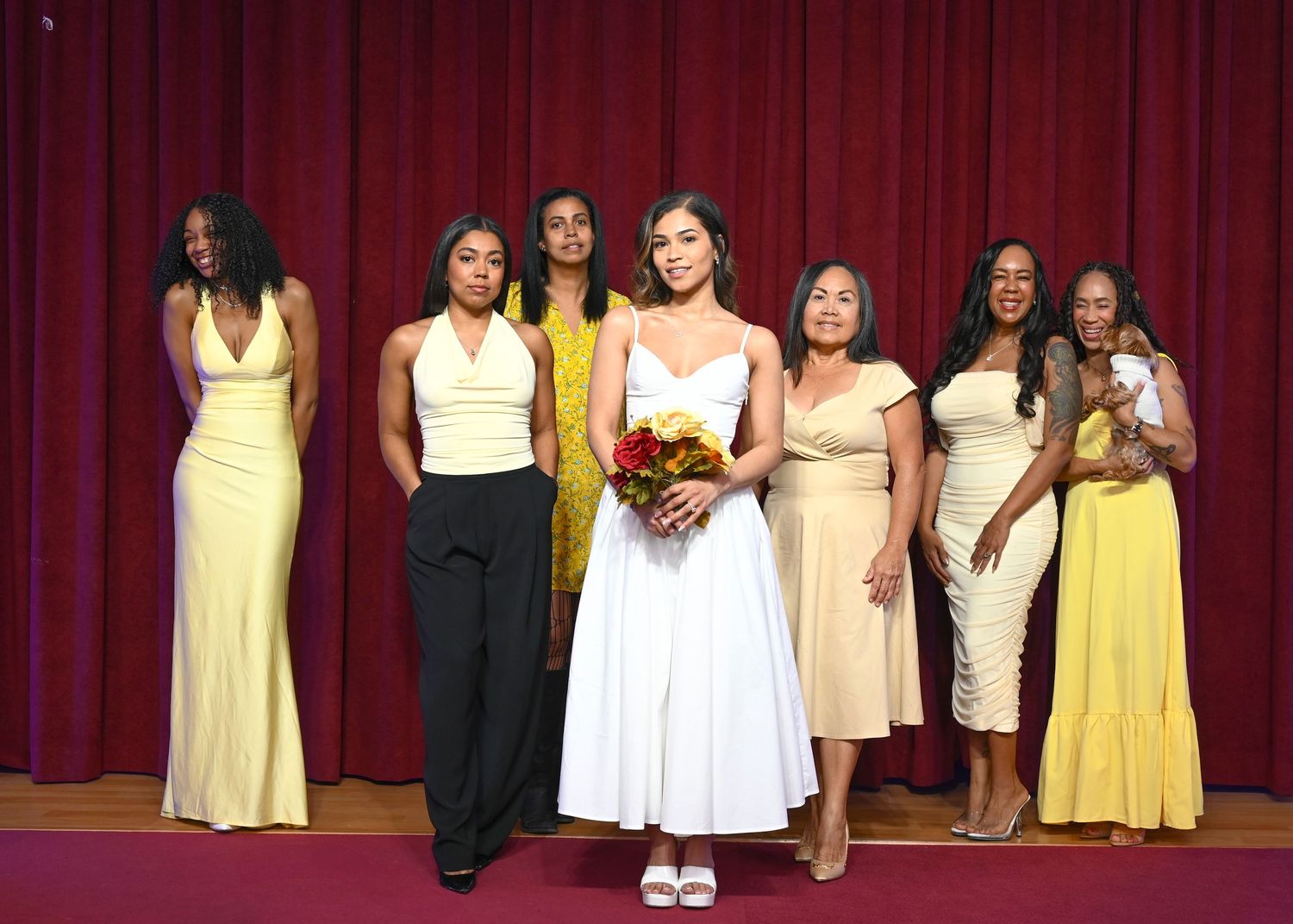 Bridesmaids in yellow and neutral dresses pose with bride holding red bouquet on stage.