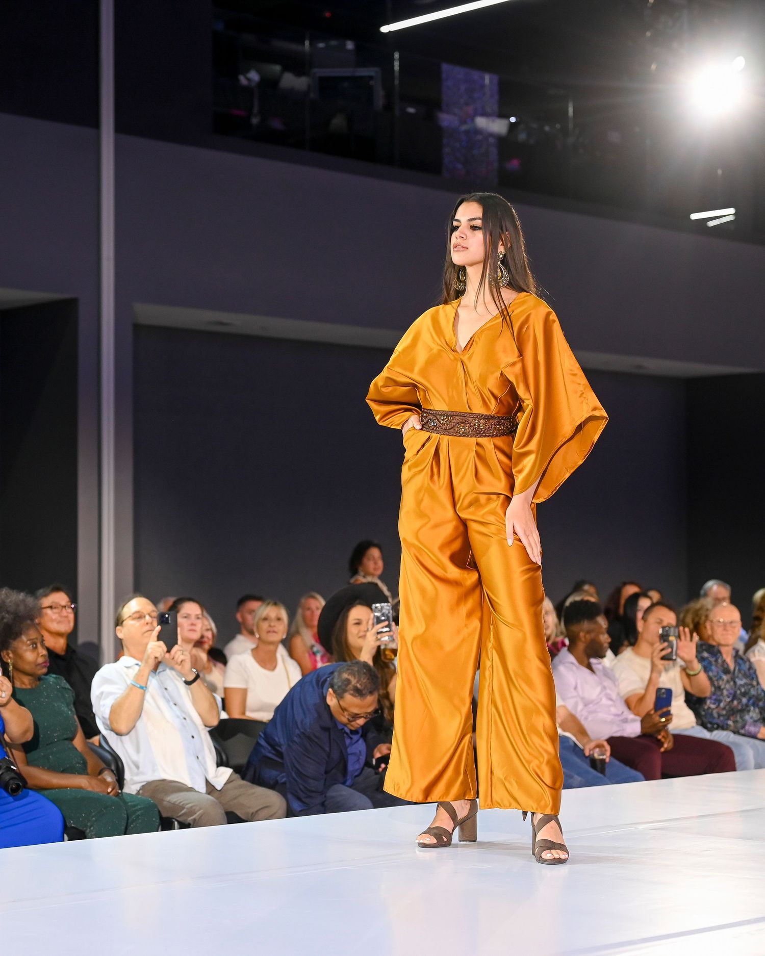 Model walks runway in flowing orange dress with seated audience.