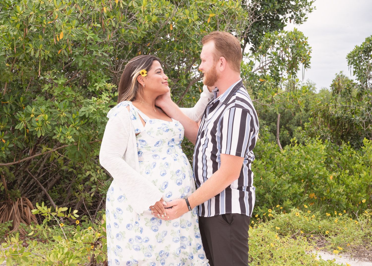 Pregnant couple sharing a tender moment outdoors, woman in floral dress with yellow flower in hair, man in striped shirt.