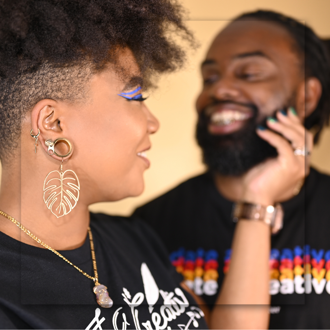 A stylish person with natural hair and leaf earrings smiles while someone talks on phone in background.