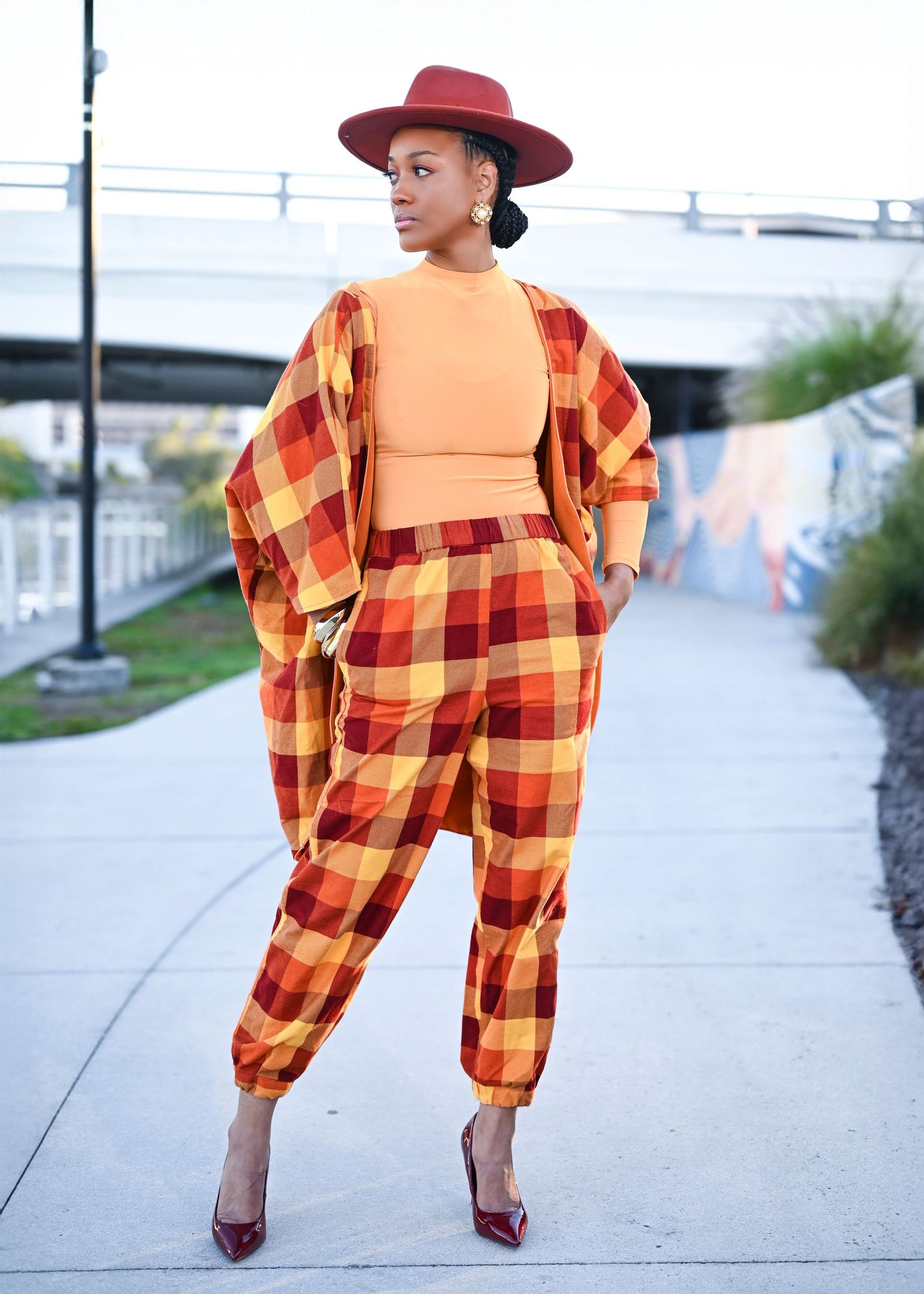 Fashion model in orange plaid coordinates and burgundy hat poses on urban path.