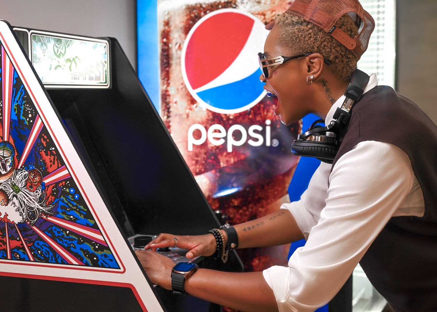 Person wearing sunglasses and headphones plays a retro arcade game cabinet in front of a Pepsi advertisement.