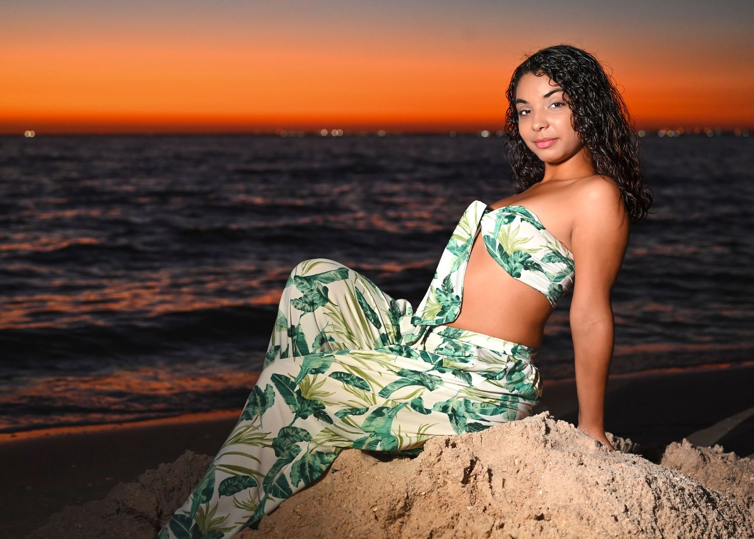 Woman with curly dark hair poses on a sandy beach in a tropical leaf-print crop top and skirt set at sunset over the ocean.