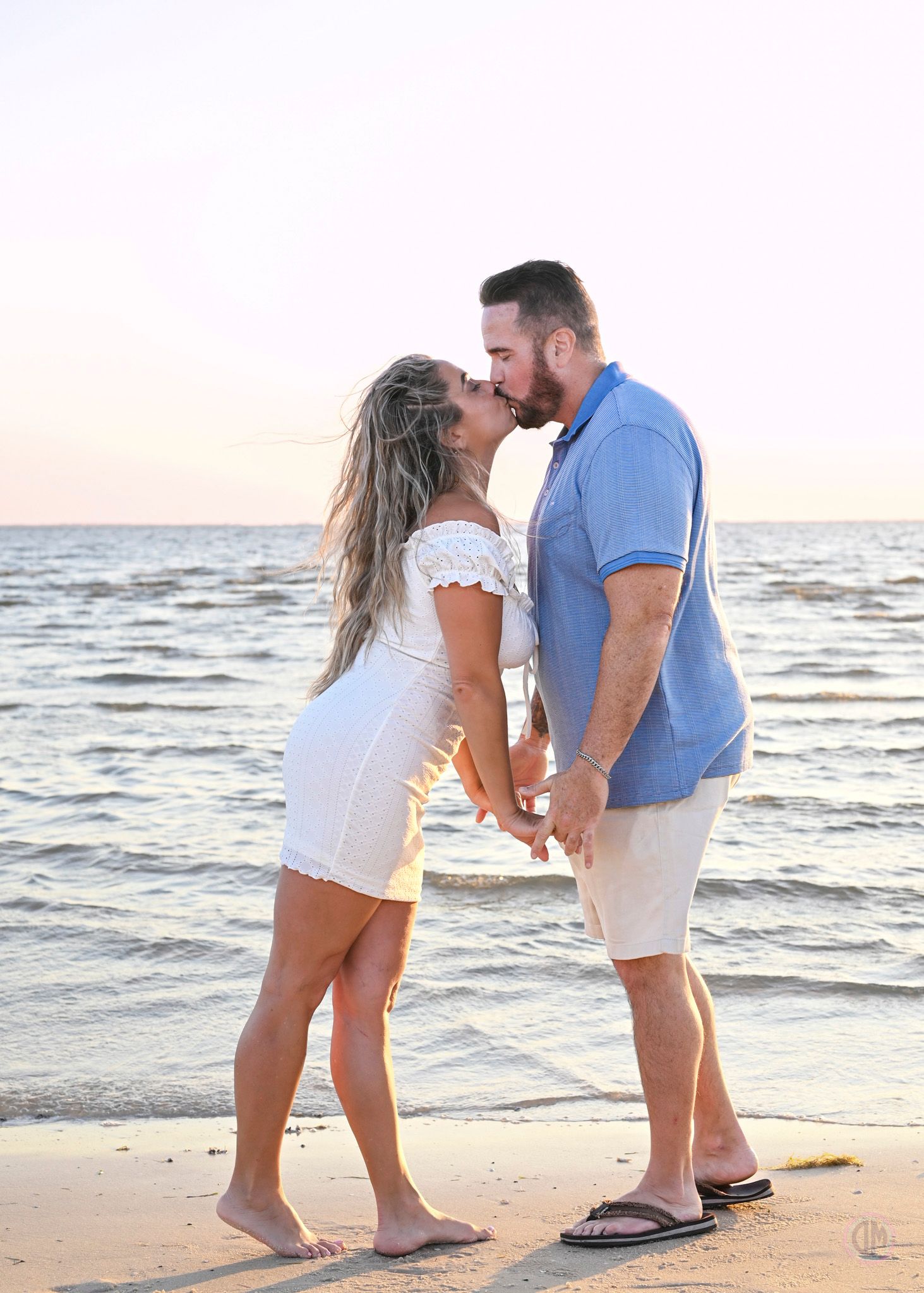 Couple kissing on beach at sunset, woman in white dress holding hands with man in blue polo shirt.