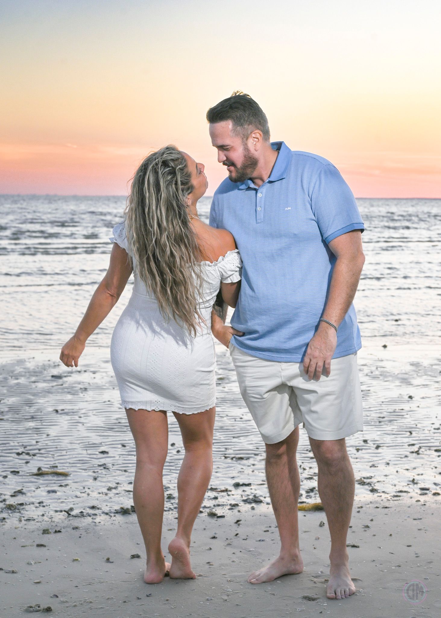 Couple laughing together on a beach at sunset, woman in white dress and man in blue polo shirt.