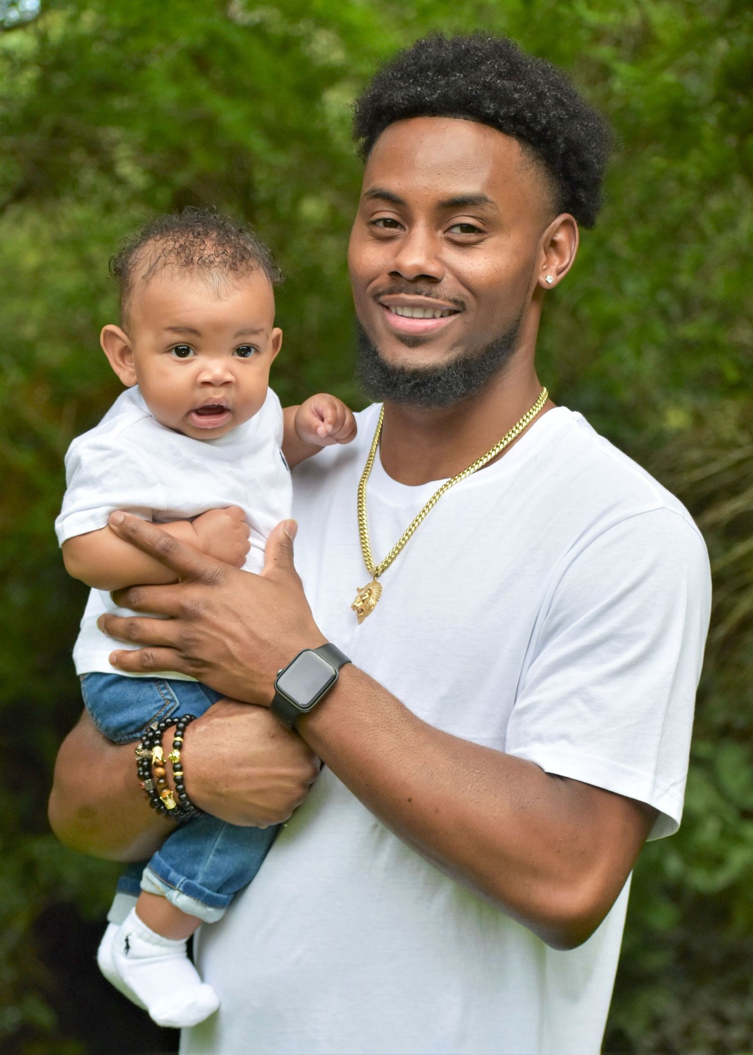 Smiling man in a white t-shirt and gold chain holds a baby in a white top and denim shorts outdoors in front of green trees.
