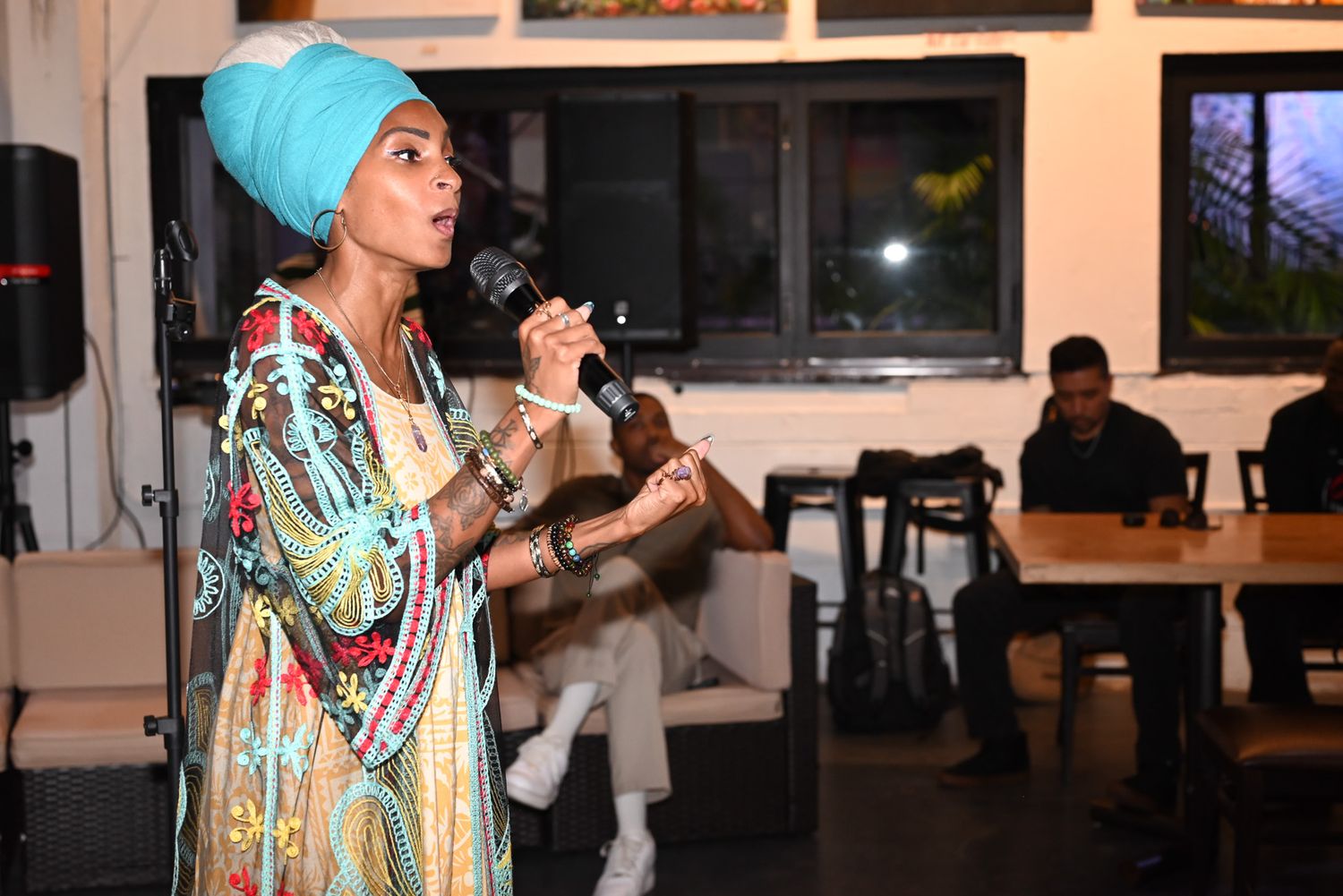 Woman in a turquoise head wrap and colorful patterned robe sings into a microphone at an indoor venue while guests sit nearby.