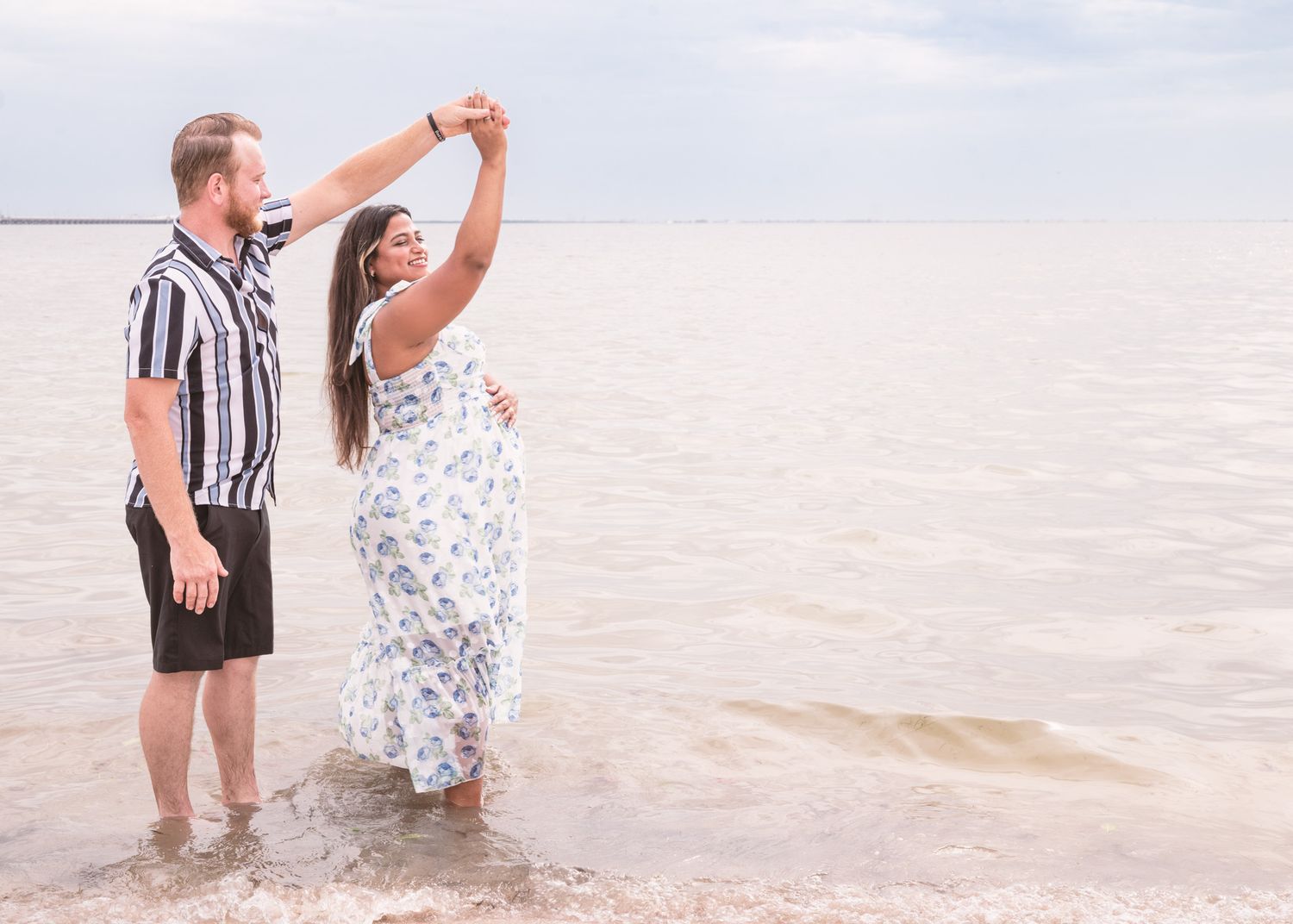 Couple dancing in shallow ocean water, man spinning woman in floral dress on cloudy beach day.